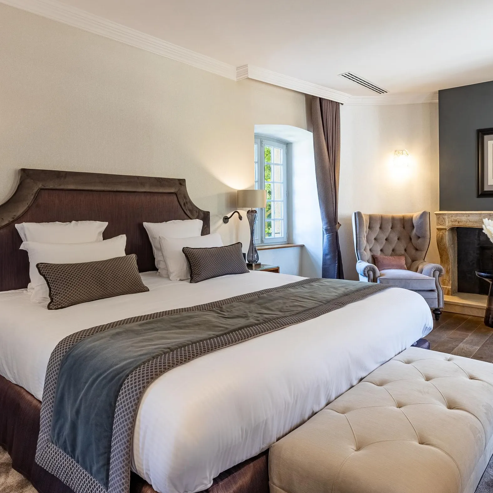 Chambre élégante avec un grand lit à tête en bois, une cheminée, des lampes murales et un fauteuil rembourré près d'une fenêtre