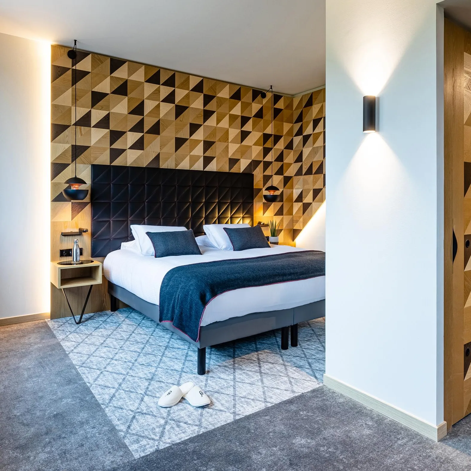 Chambre avec un lit double, une tête de lit en bois à motifs géométriques, une table de chevet et une paire de chaussons sur un tapis