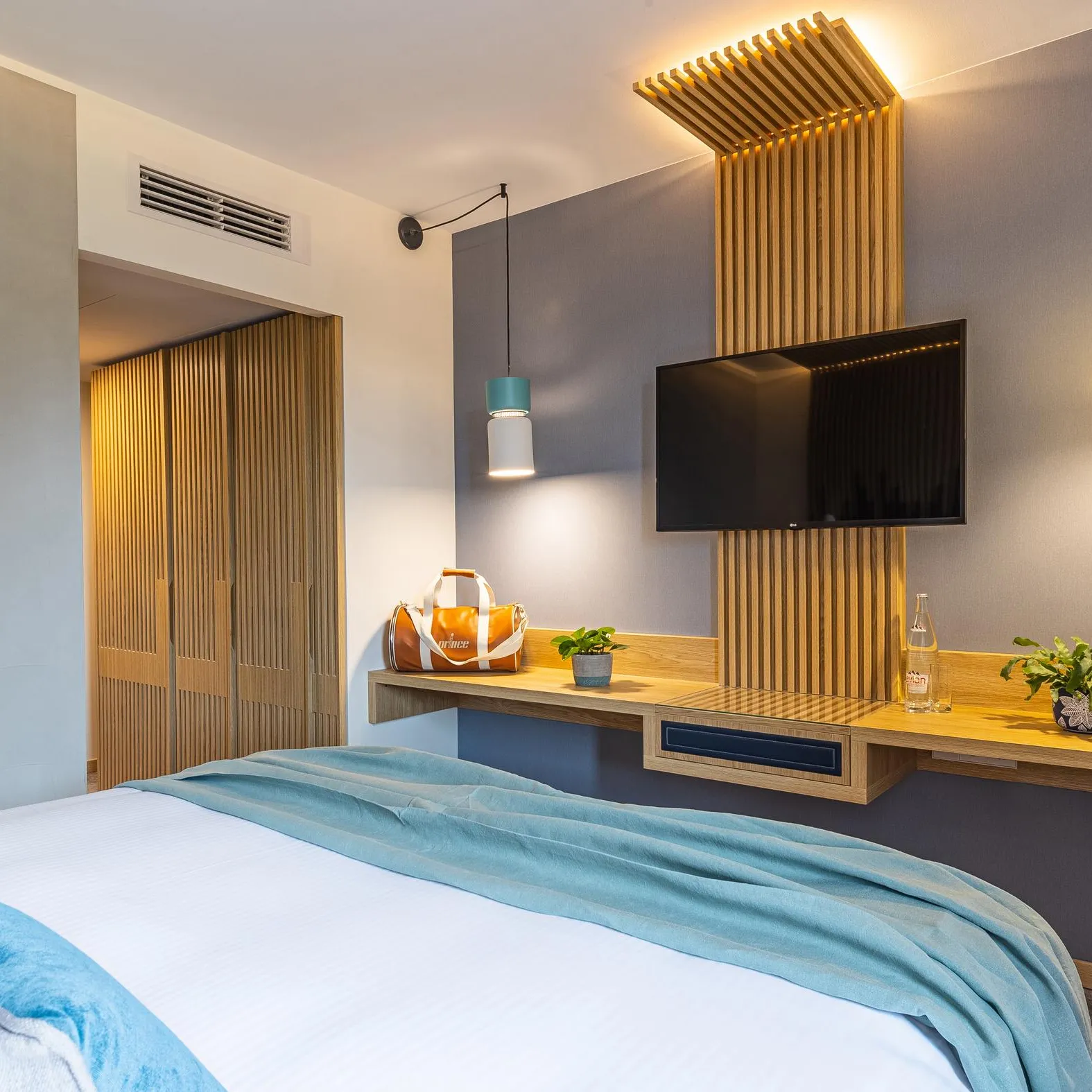 Chambre moderne avec un lit recouvert d'une couverture bleue, une télévision montée sur le mur et une table de chevet en bois avec divers objets