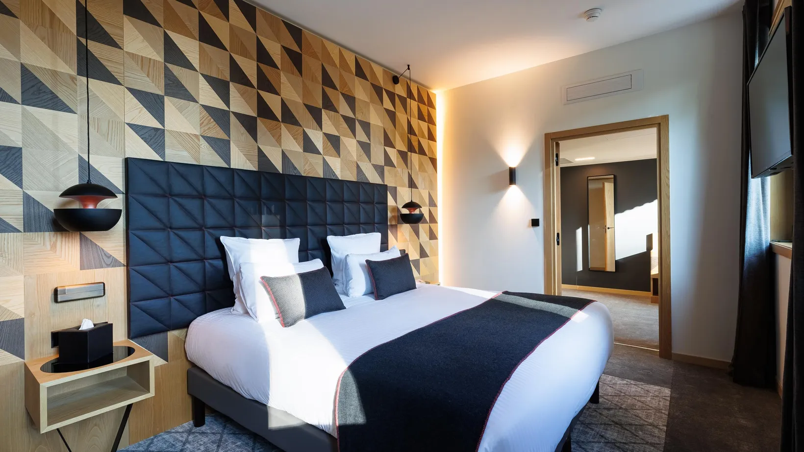 Modernes Zimmer mit einem Doppelbett, einem Kopfteil mit geometrischen Mustern und einer mit dreieckigen Holzpaneelen in verschiedenen Farbtönen verzierten Wand
