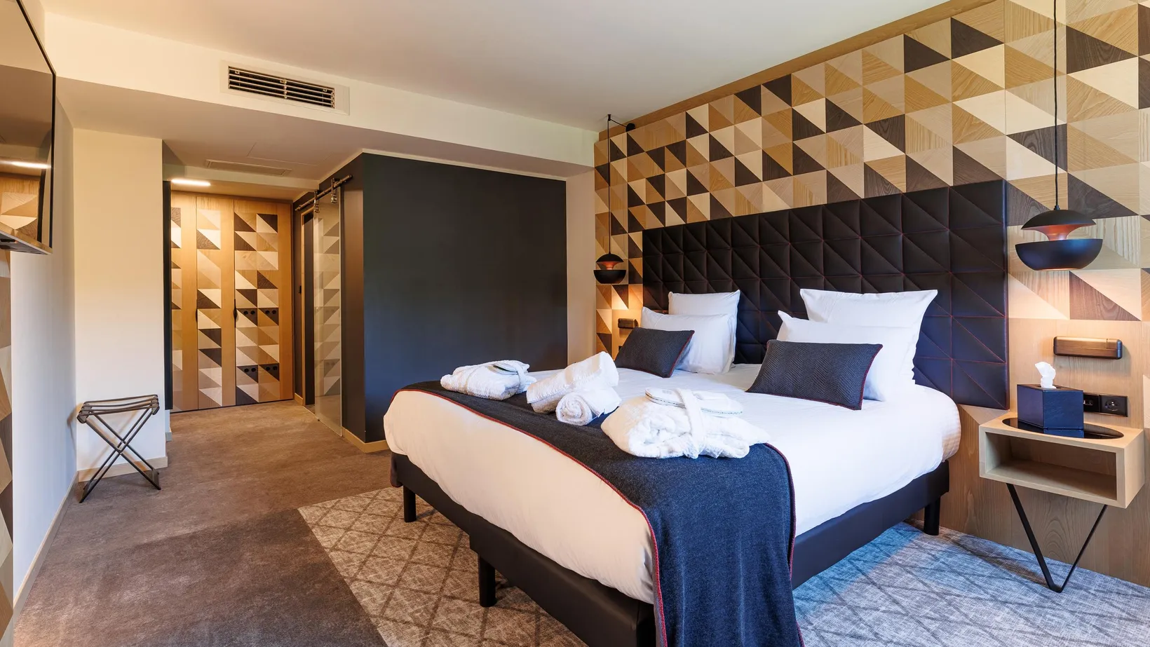 Modernes Hotelzimmer mit Doppelbett, geometrischem Holzkopfteil, Designleuchten und offener Tür zu einem ähnlichen Flur