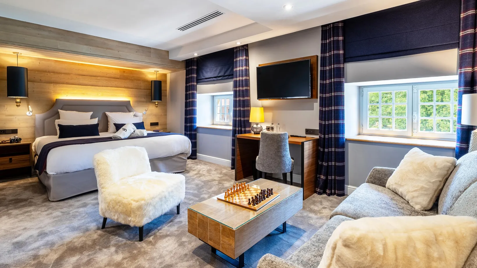 Spacious room with bed, sofa, armchair, coffee table with chessboard, desk, and television, decorated in gray and blue tones with wooden elements and windows.
