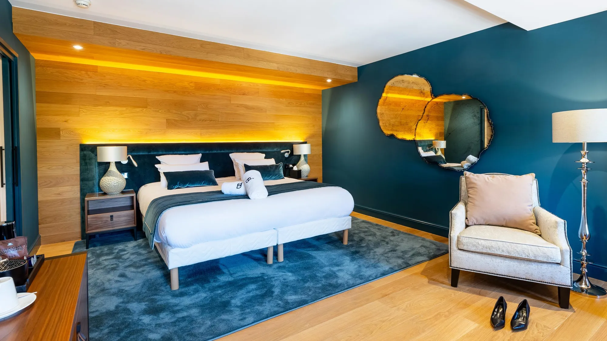 Chambre avec lit double, miroir rond, fauteuil et table de chevet, murs bleu foncé et bois clair, éclairage doux