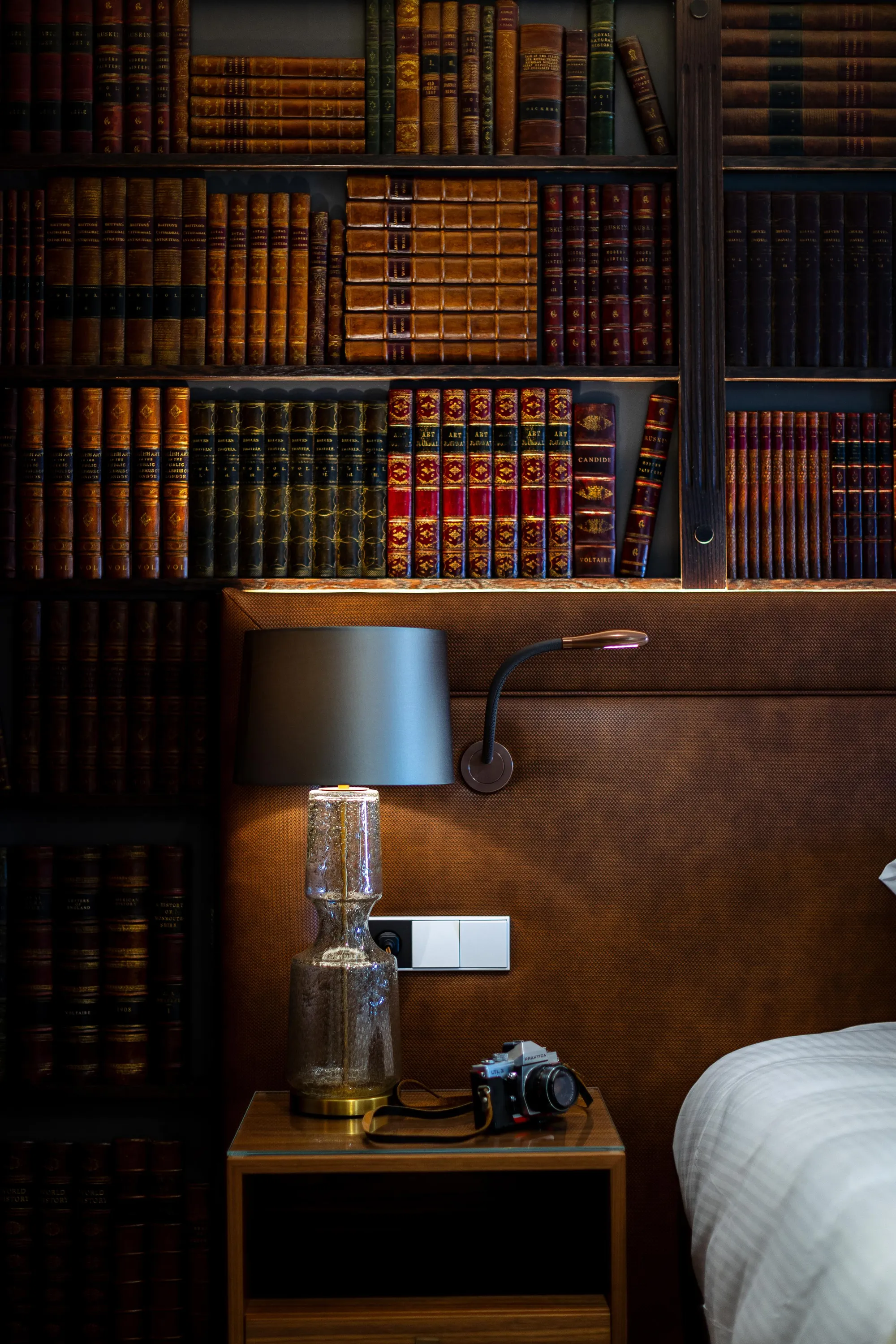 Une table de chevet avec une lampe, un vase et un appareil photo devant une bibliothèque remplie de livres