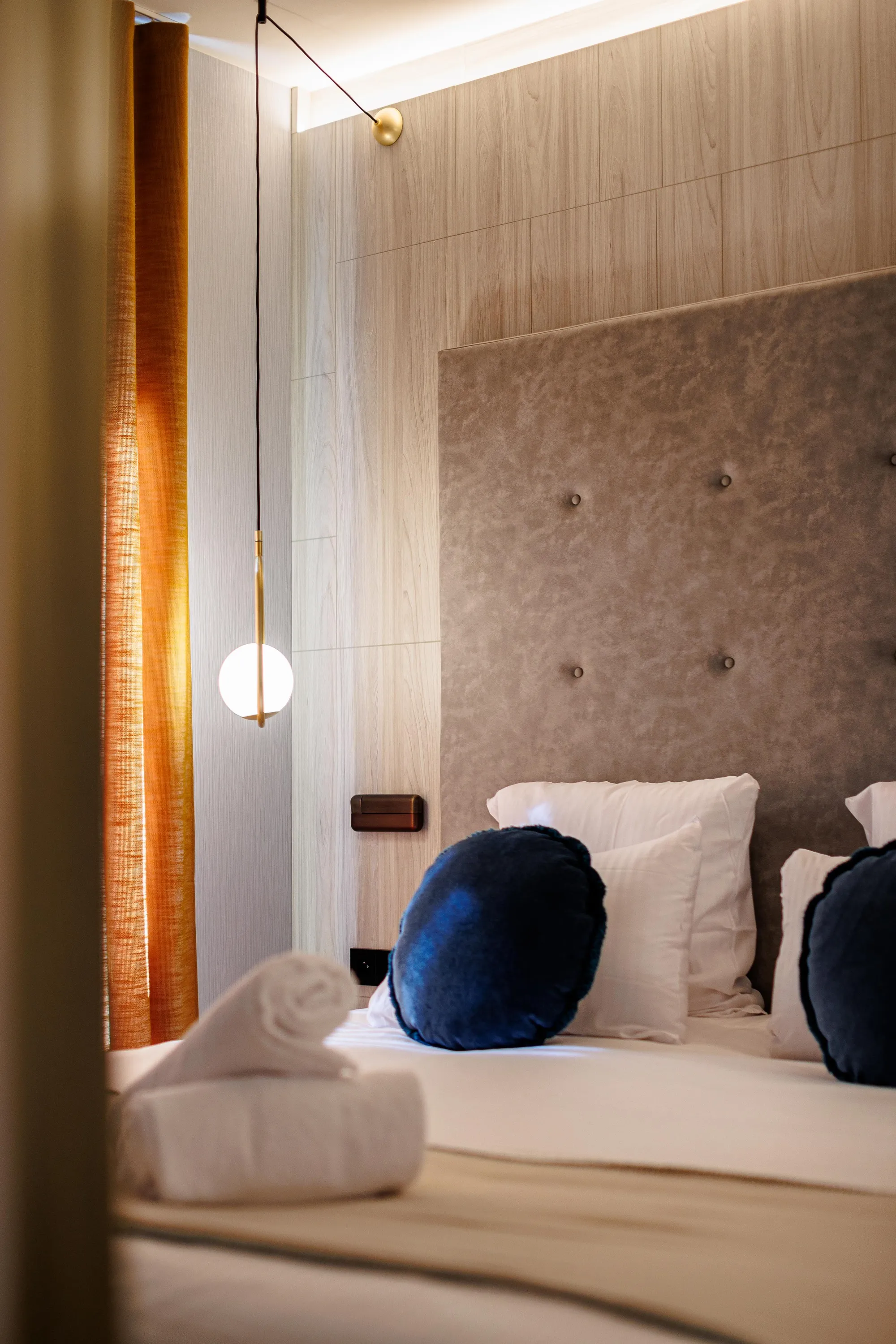 Bett mit grauer Stoffkopflehne und Kissen, einer Hängelampe und einem Fenster mit orangefarbenem Vorhang
