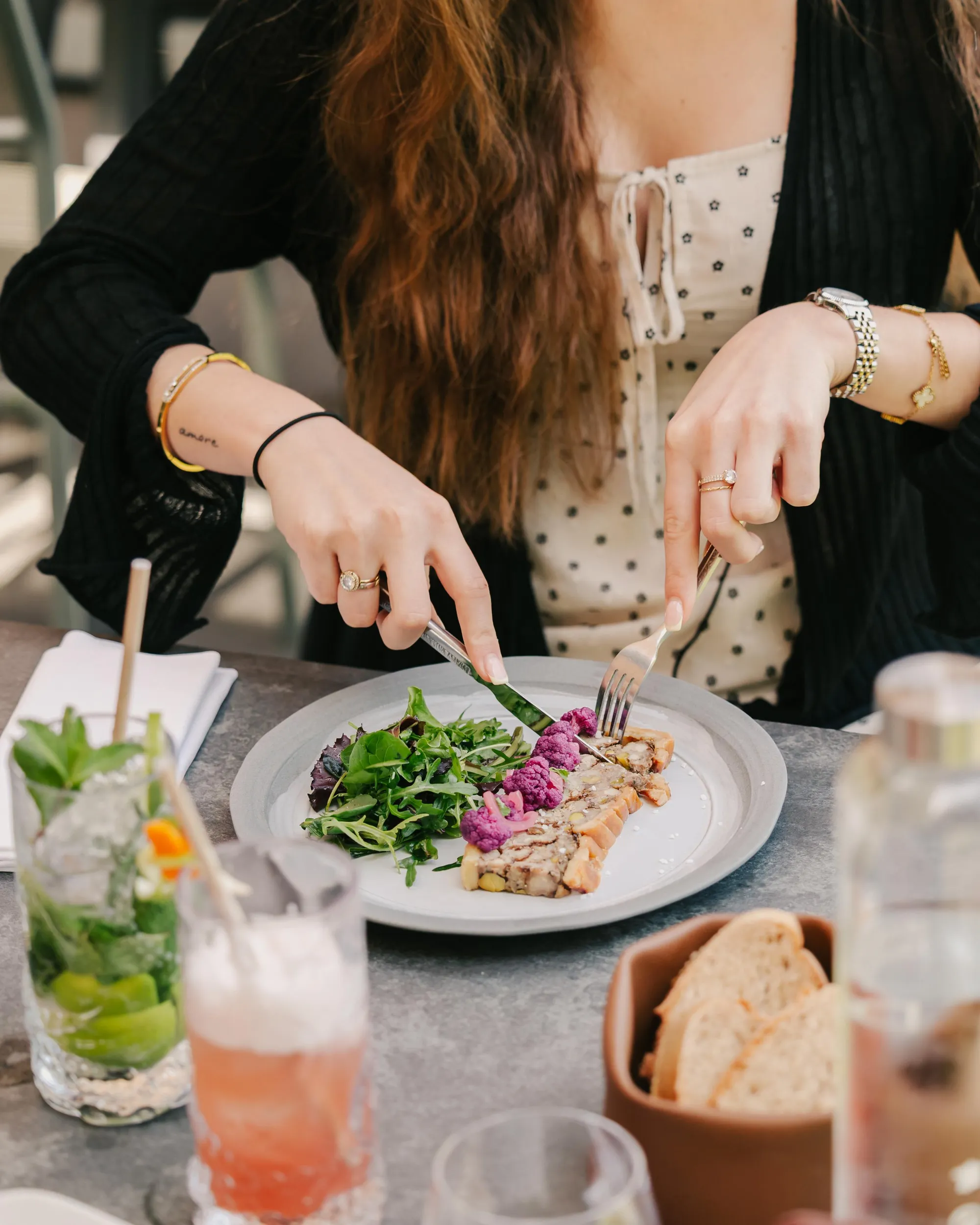 Zwei Personen verwenden Besteck, um eine Schüssel mit Fleisch, Gemüse und Gurken zu essen, begleitet von Brot und Getränken auf einem Tisch.