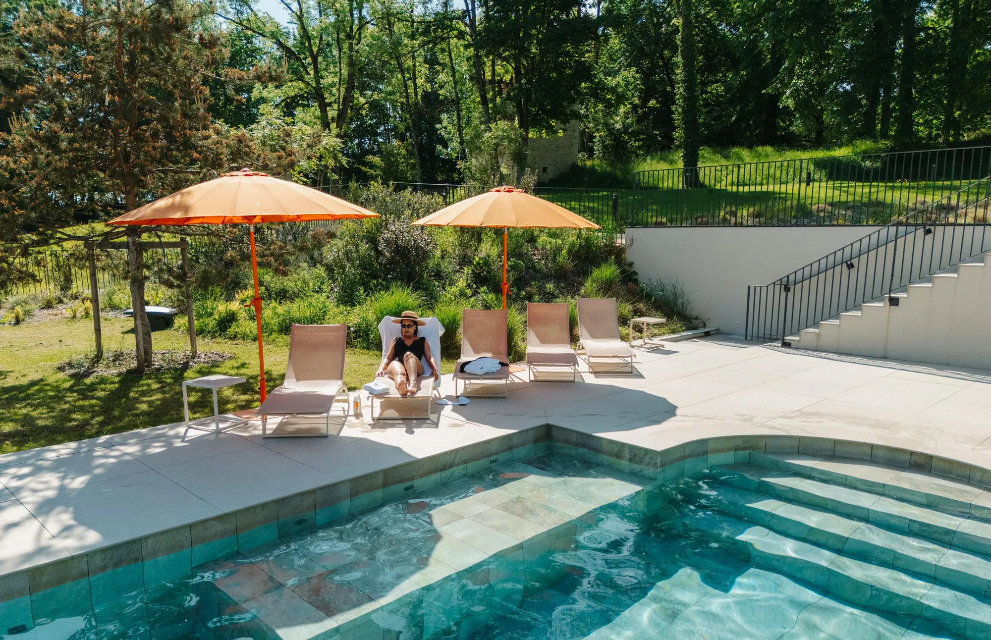 Une personne est assise sur un transat près d'une piscine, entourée de chaises longues et de parasols orange, dans un jardin arboré