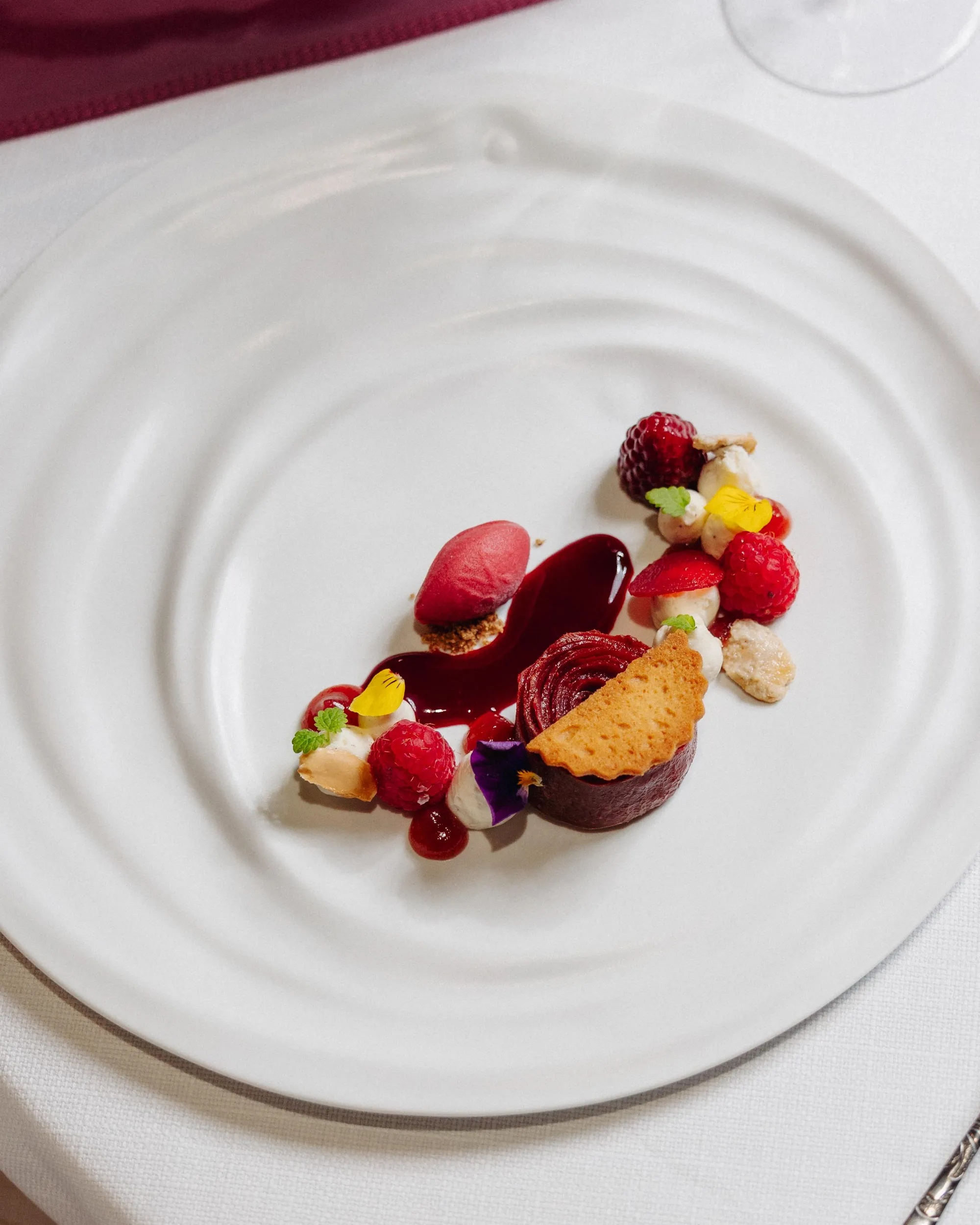 Assiette blanche avec une composition de dessert à base de fruits rouges, de crème, de biscuit et de sauce, présentée sur une nappe blanche