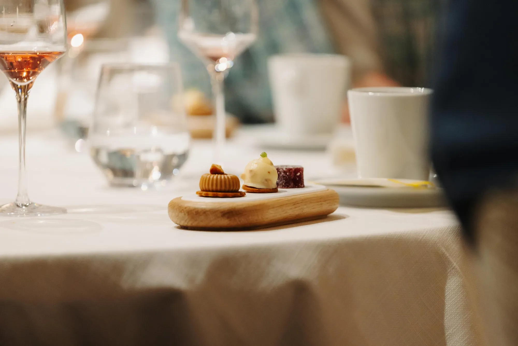 Kleine Holzschale mit drei Desserts und Weingläsern im Hintergrund auf einem mit einer weißen Tischdecke bedeckten Tisch