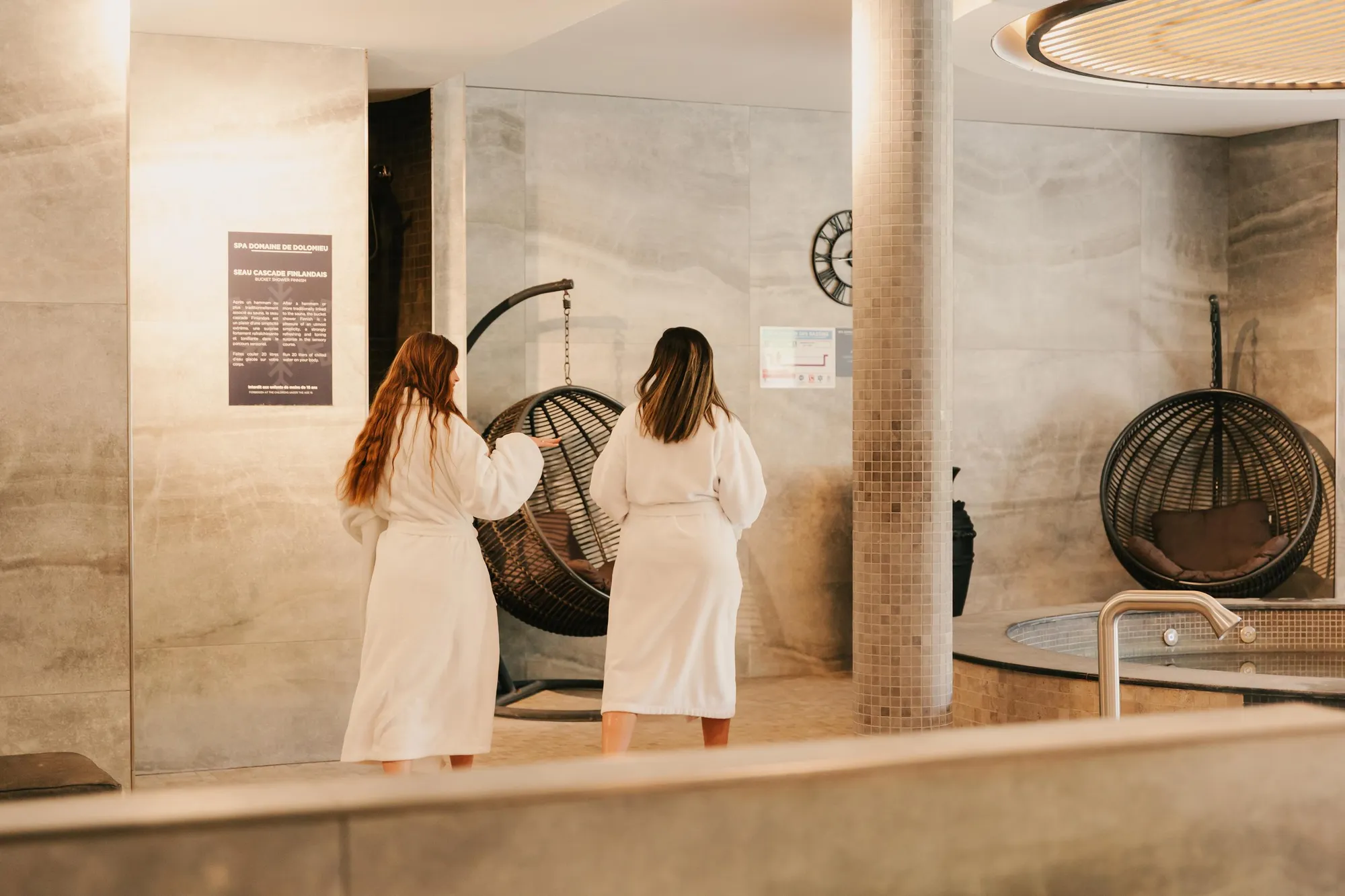 Two people in white swimsuits stand in front of a glass door in a spa, with information displayed on a sign to the left.