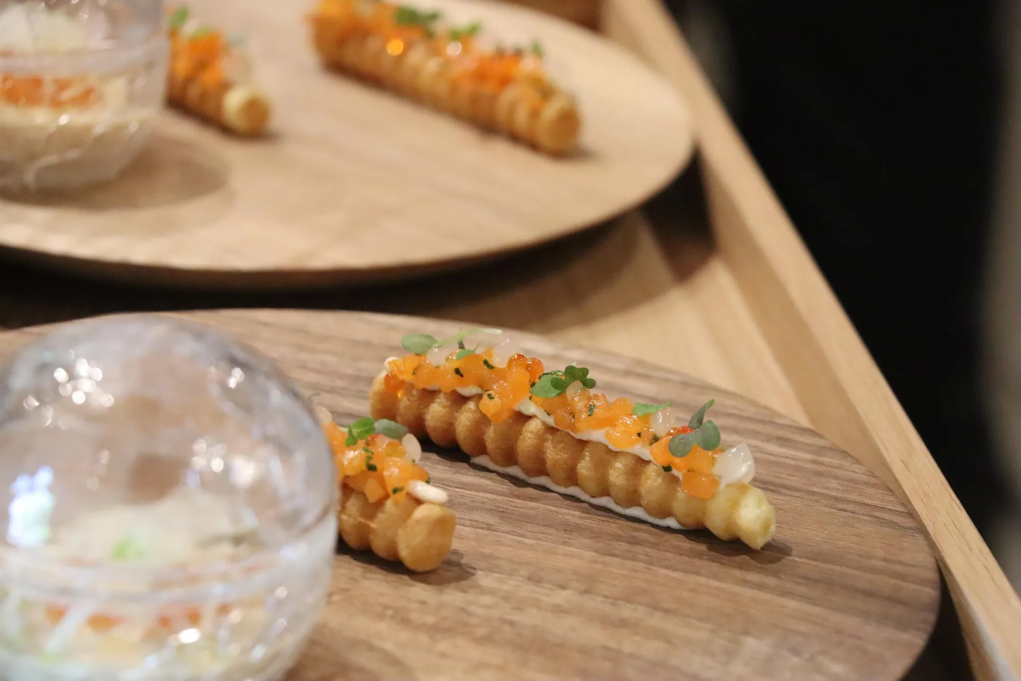 Un plat composé de petits gâteaux recouverts de caviar et de micro-pousses, présenté sur une assiette en bois avec un verre transparent à côté