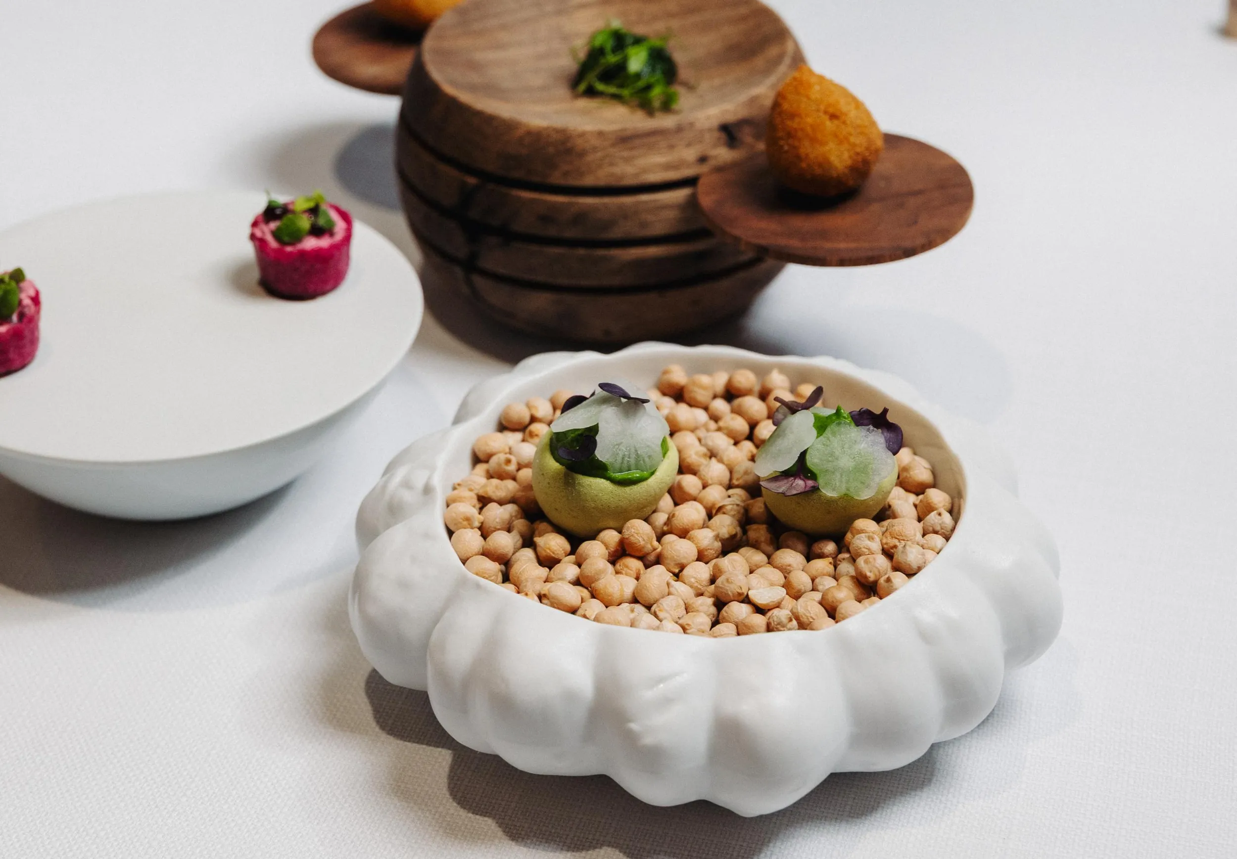 Un plat blanc rempli de pois chiches avec deux boules vertes garnies de fines herbes et de tranches de légumes, posé sur une table blanche
