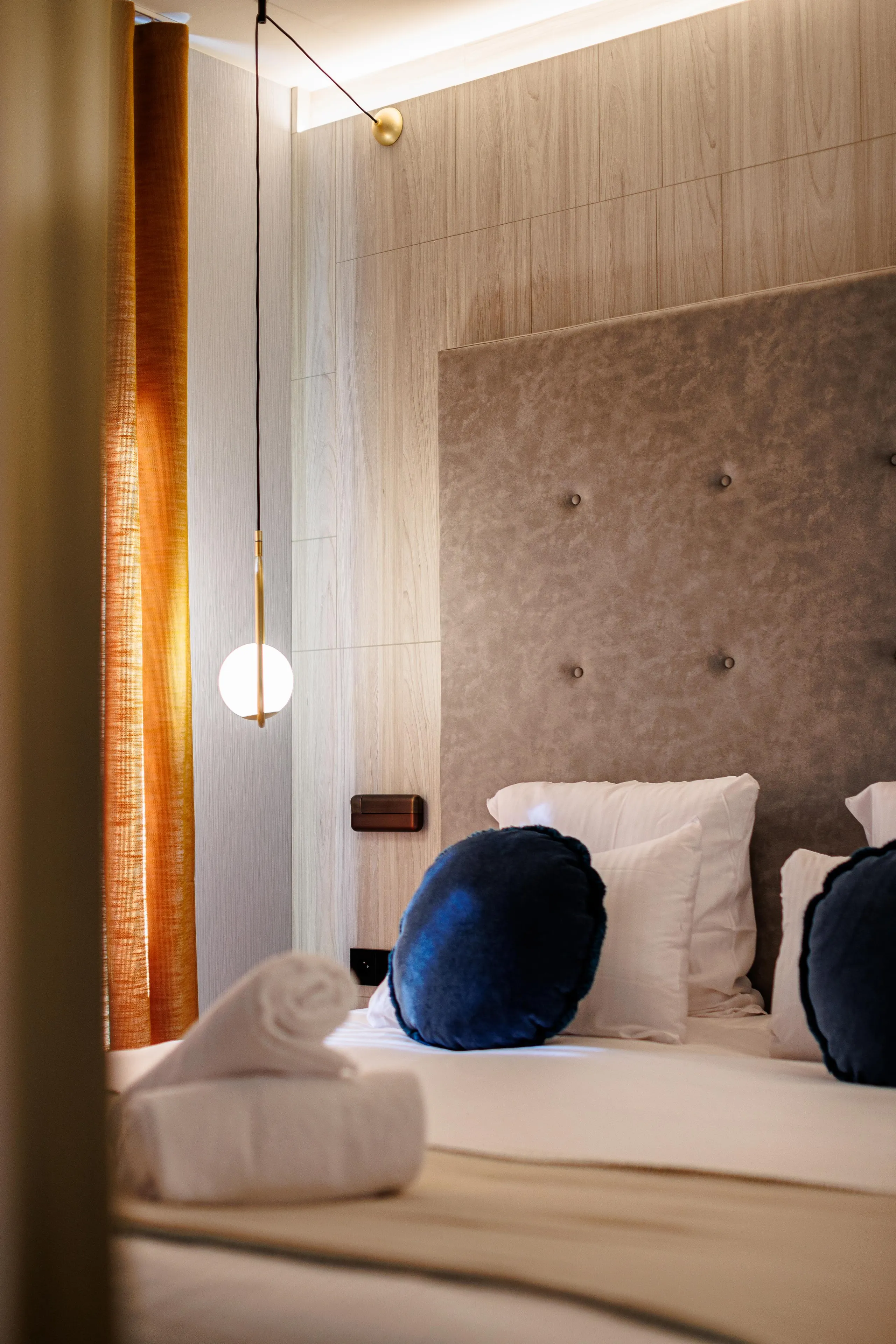 Lit avec une tête de lit en tissu gris et des coussins, une lampe suspendue et une fenêtre avec un rideau orange