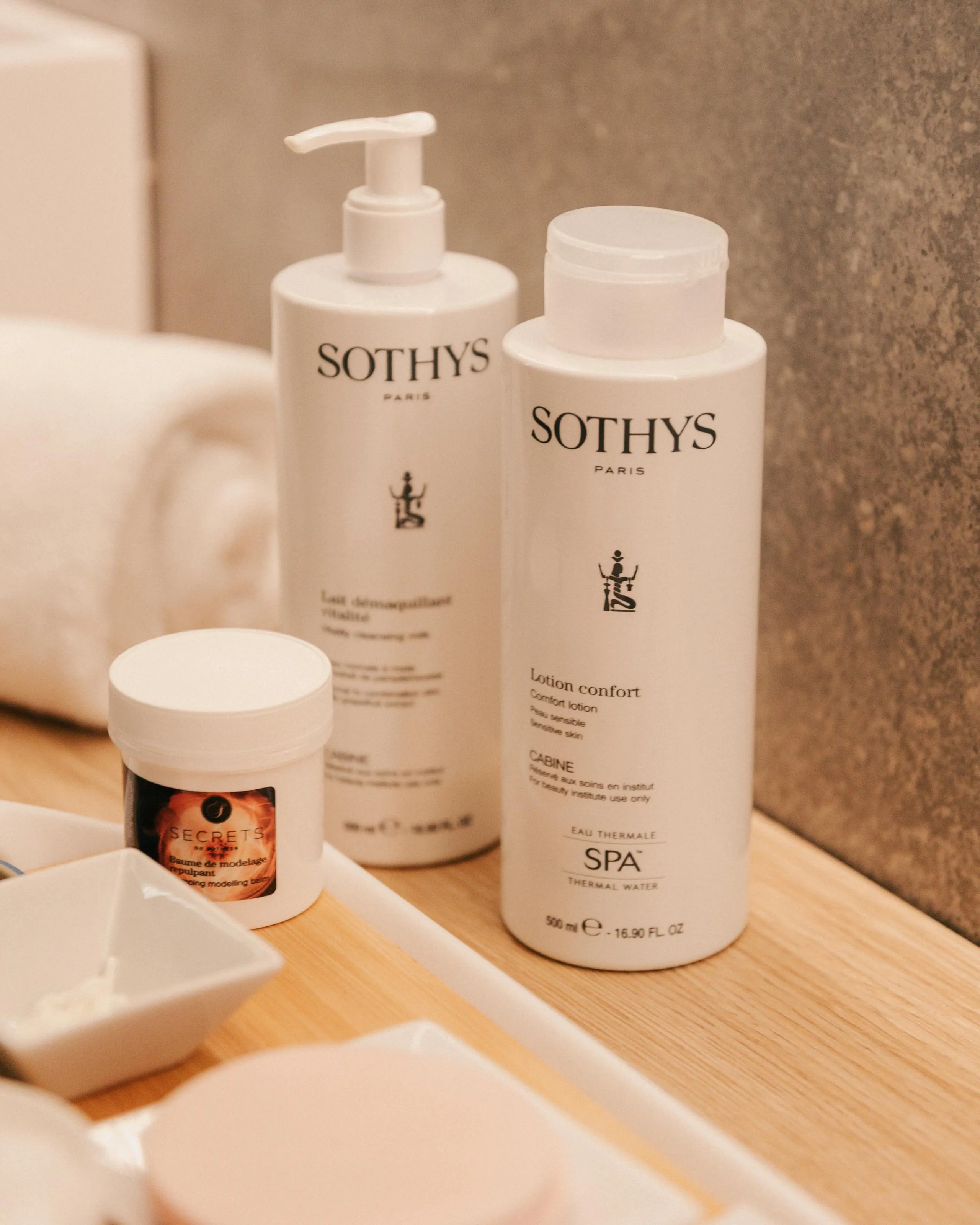 Deux flacons de produits cosmétiques Sothys Paris posés sur une surface en bois clair avec un pot de crème et une coupelle blanche
