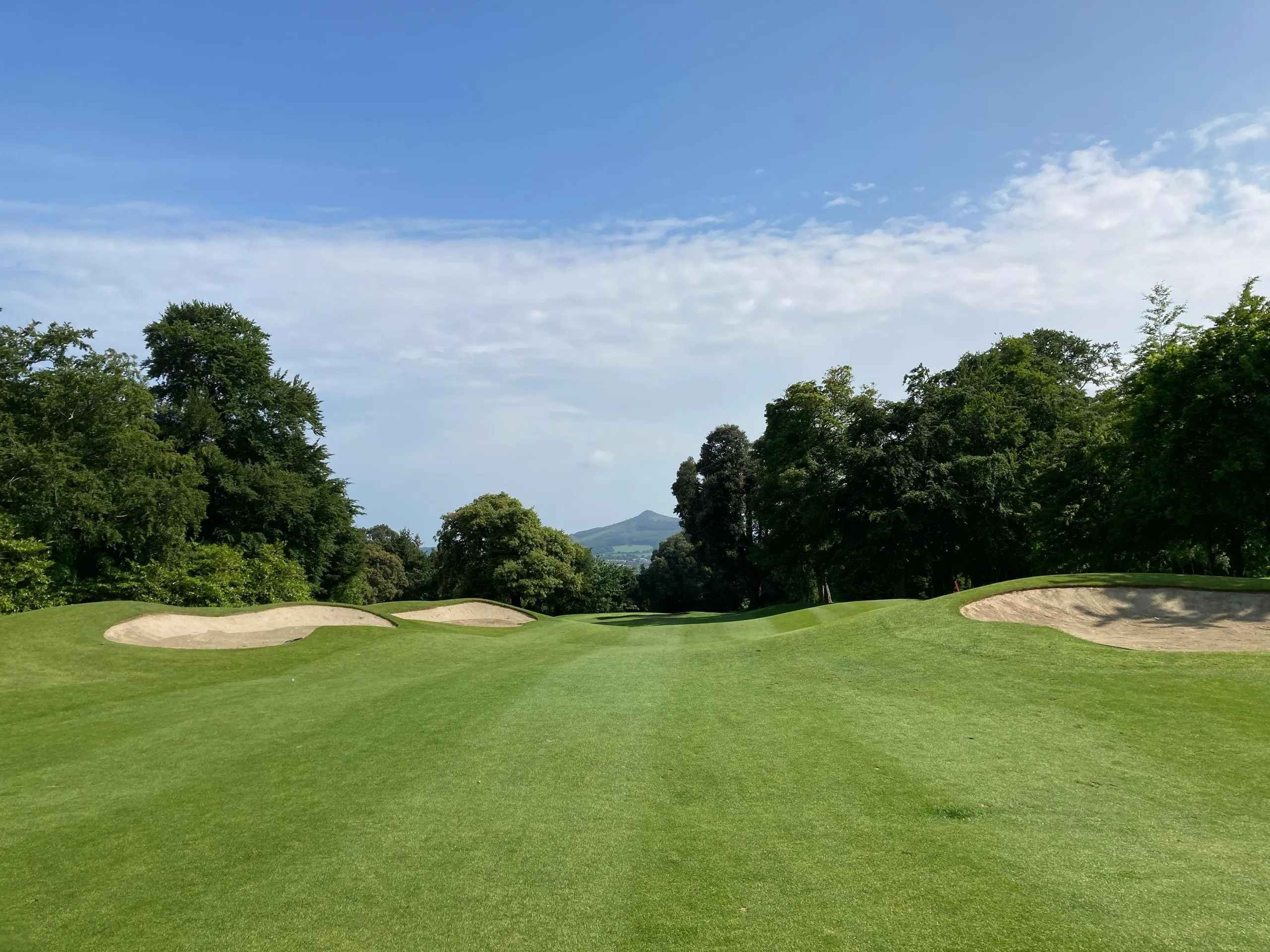 Ein Golfplatz mit Sandbunkern, umgeben von Bäumen und Hügeln unter einem teilweise bewölkten Himmel