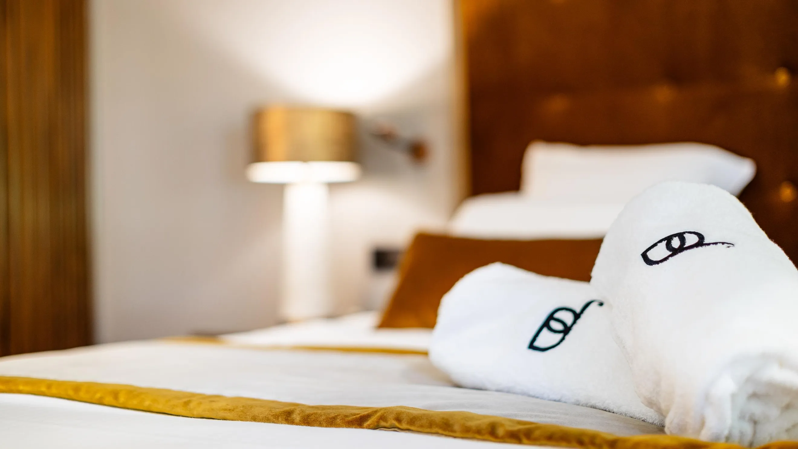Deux serviettes de bain blanches avec un logo imprimé sont posées sur un lit aux draps blancs et jaunes, avec une lampe de chevet floue en arrière-plan
