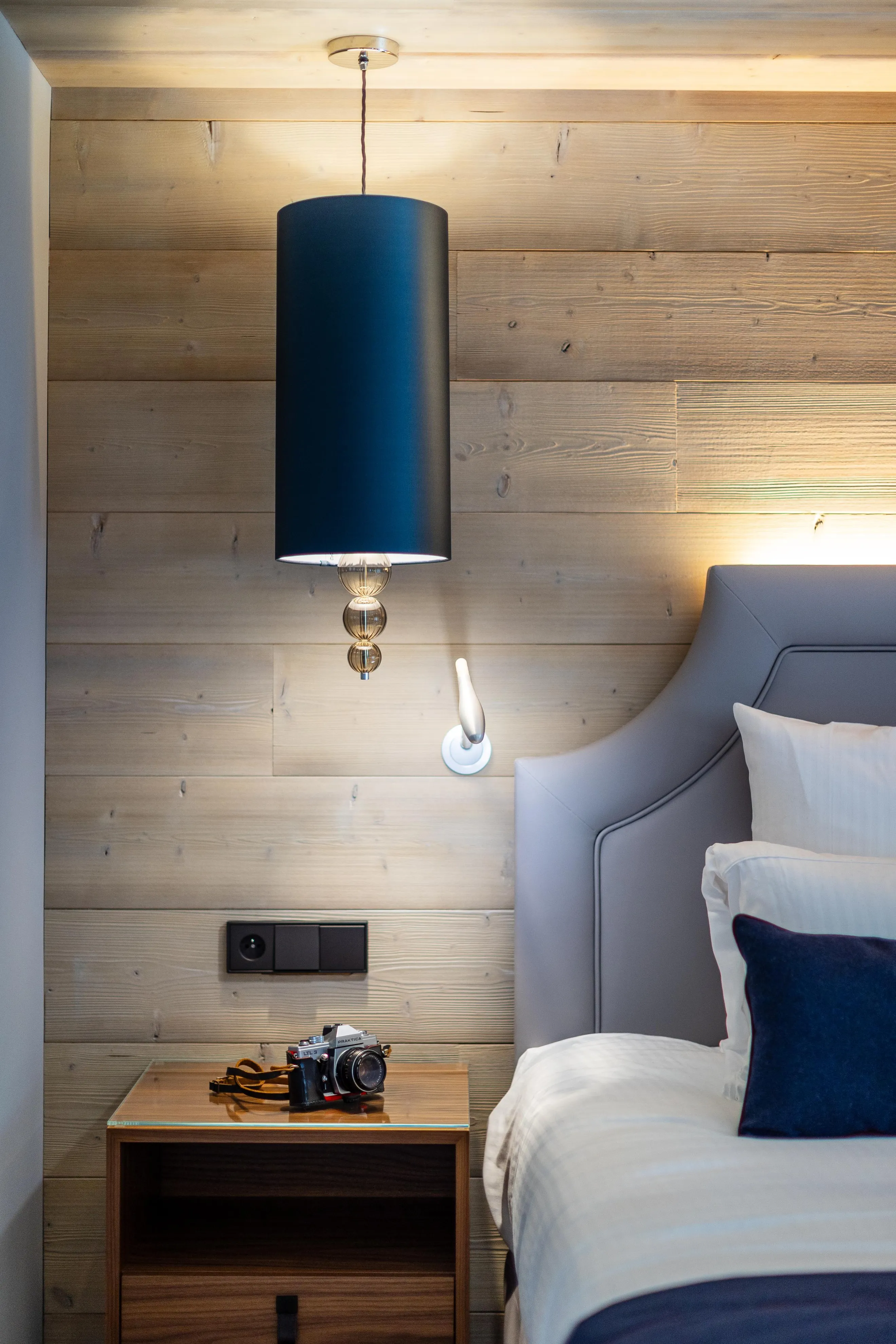 Ein Zimmer mit einem Bett, einer Hängelampe, einem Nachttisch mit einer Kamera und einer Wandlampe