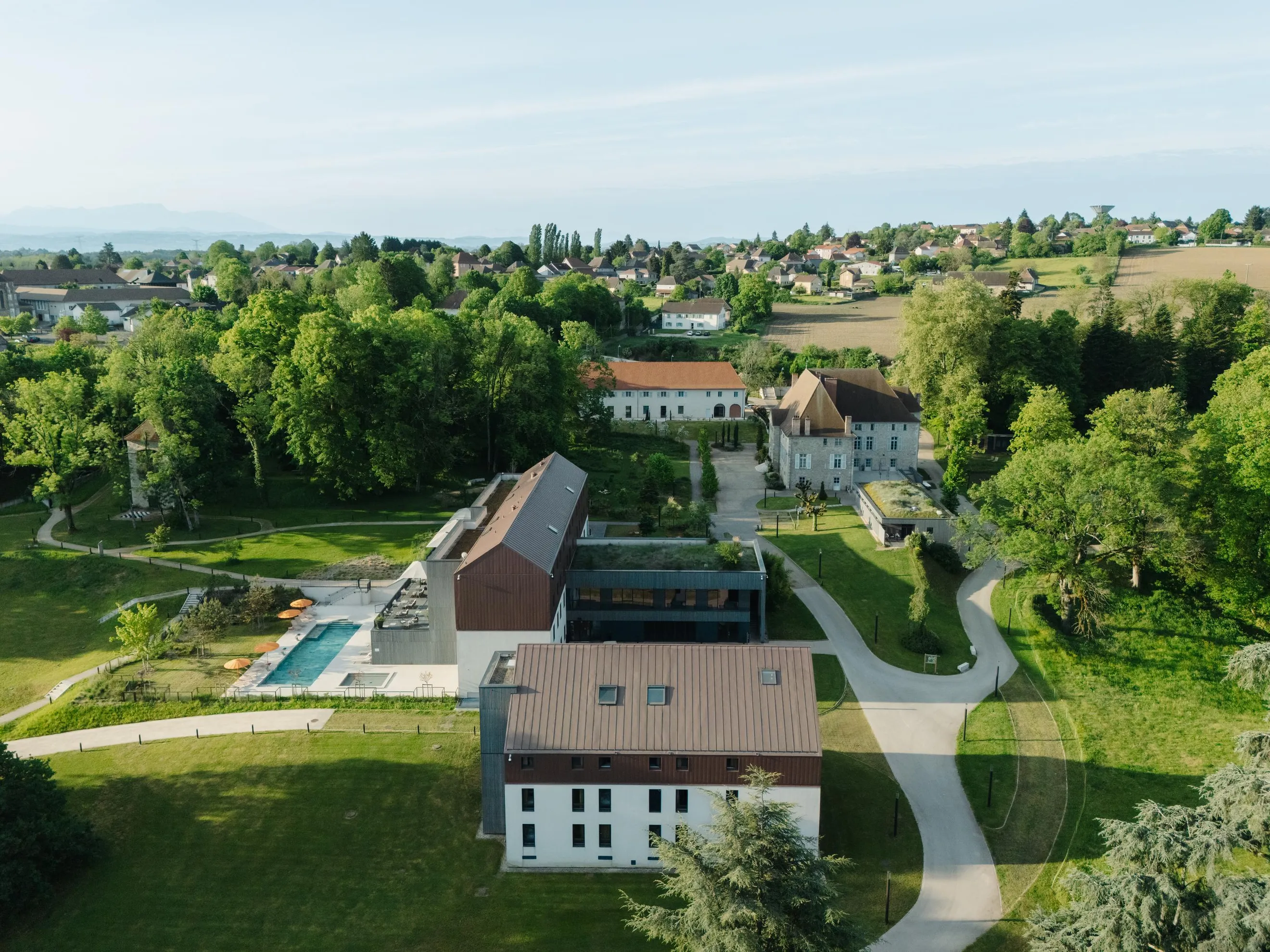 Vue aérienne d'un domaine avec bâtiments, piscines et jardins entourés d'arbres et de sentiers, en bordure d'un village et de champs
