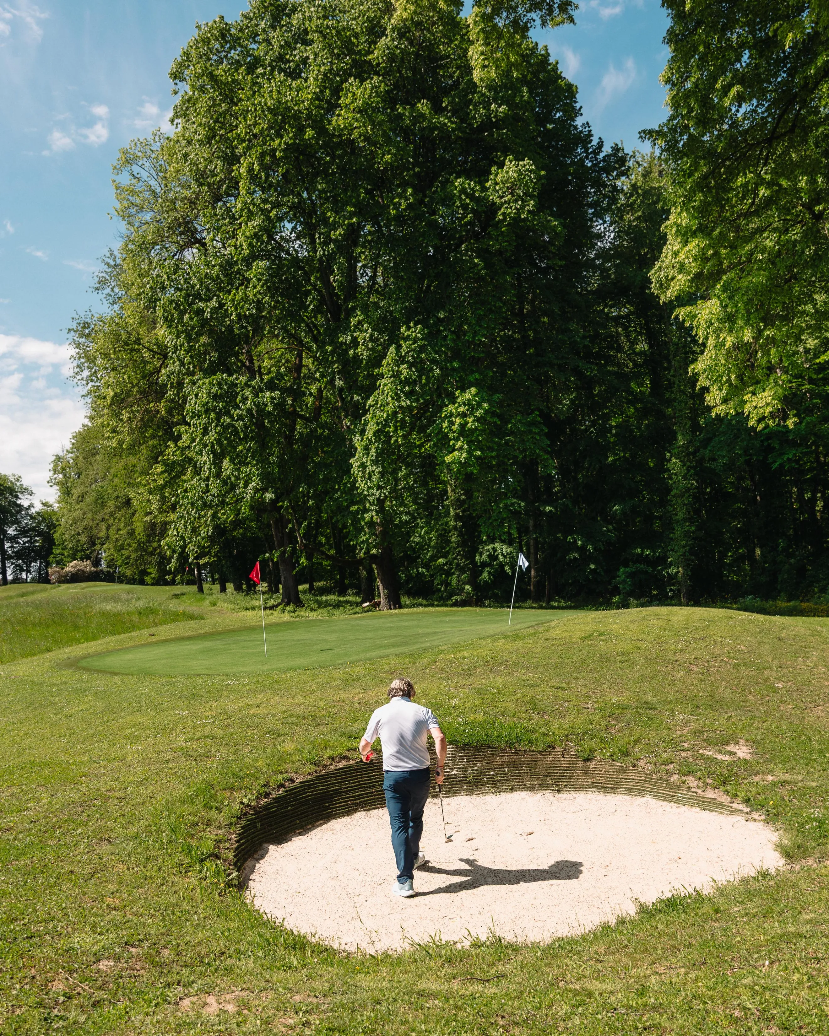 Un golfeur se tient sur un bunker de sable et regarde en direction du green entouré d'arbres