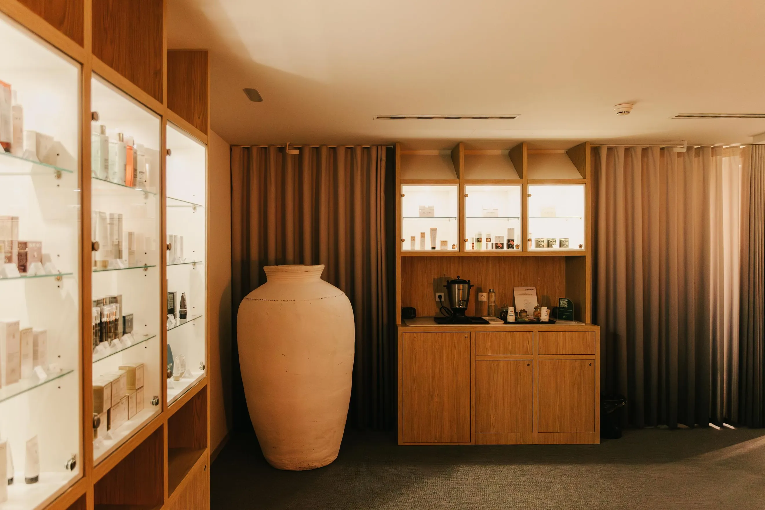 Un grand vase en terre cuite est placé devant un meuble en bois avec des étagères vitrées remplies de produits et un comptoir avec divers objets