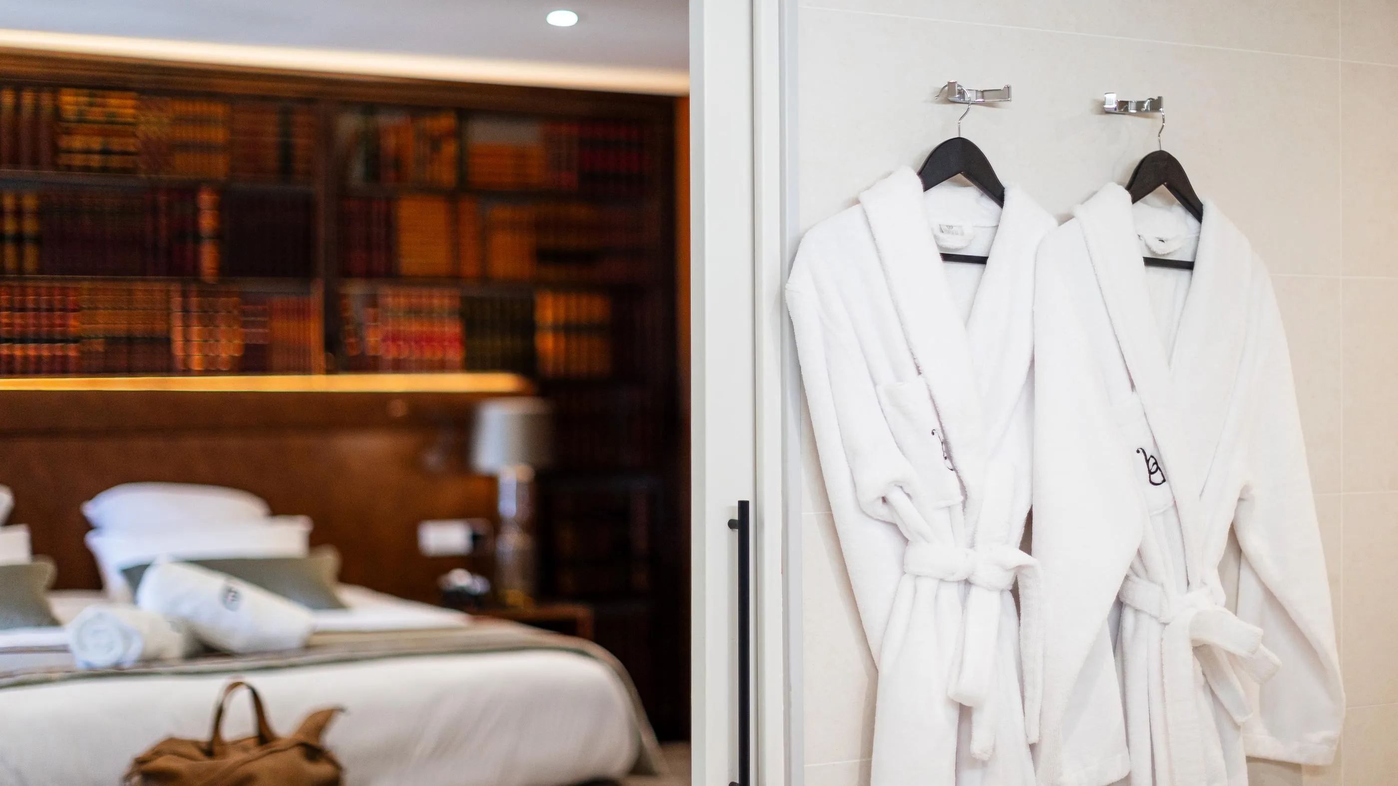 Deux peignoirs blancs suspendus à des cintres sur une porte avec en arrière-plan une chambre aux tons chauds et un lit défait