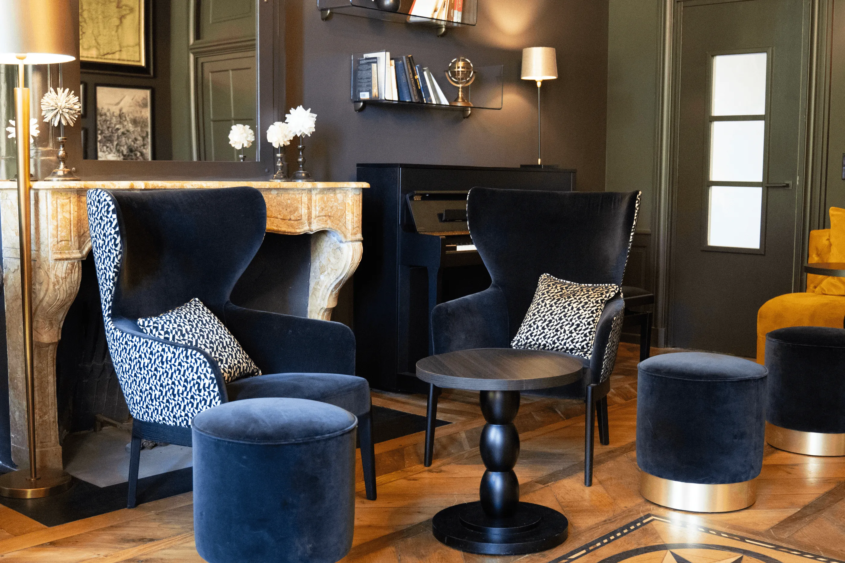 Salon avec des fauteuils et des ottomans en velours, une cheminée et des étagères avec des livres et des fleurs