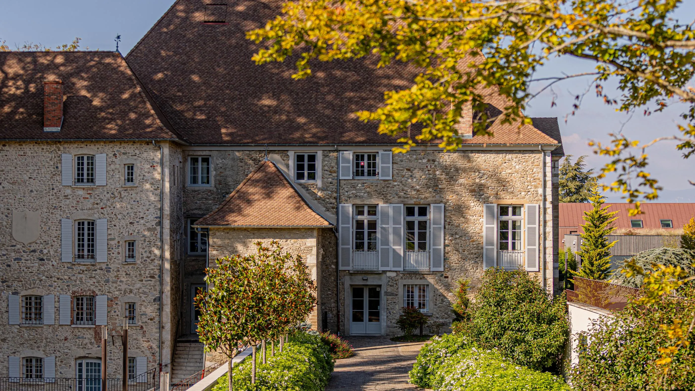 Ancienne maison en pierre avec toits en tuiles et volets gris, entourée de végétation et d'arbres, vue depuis un jardin