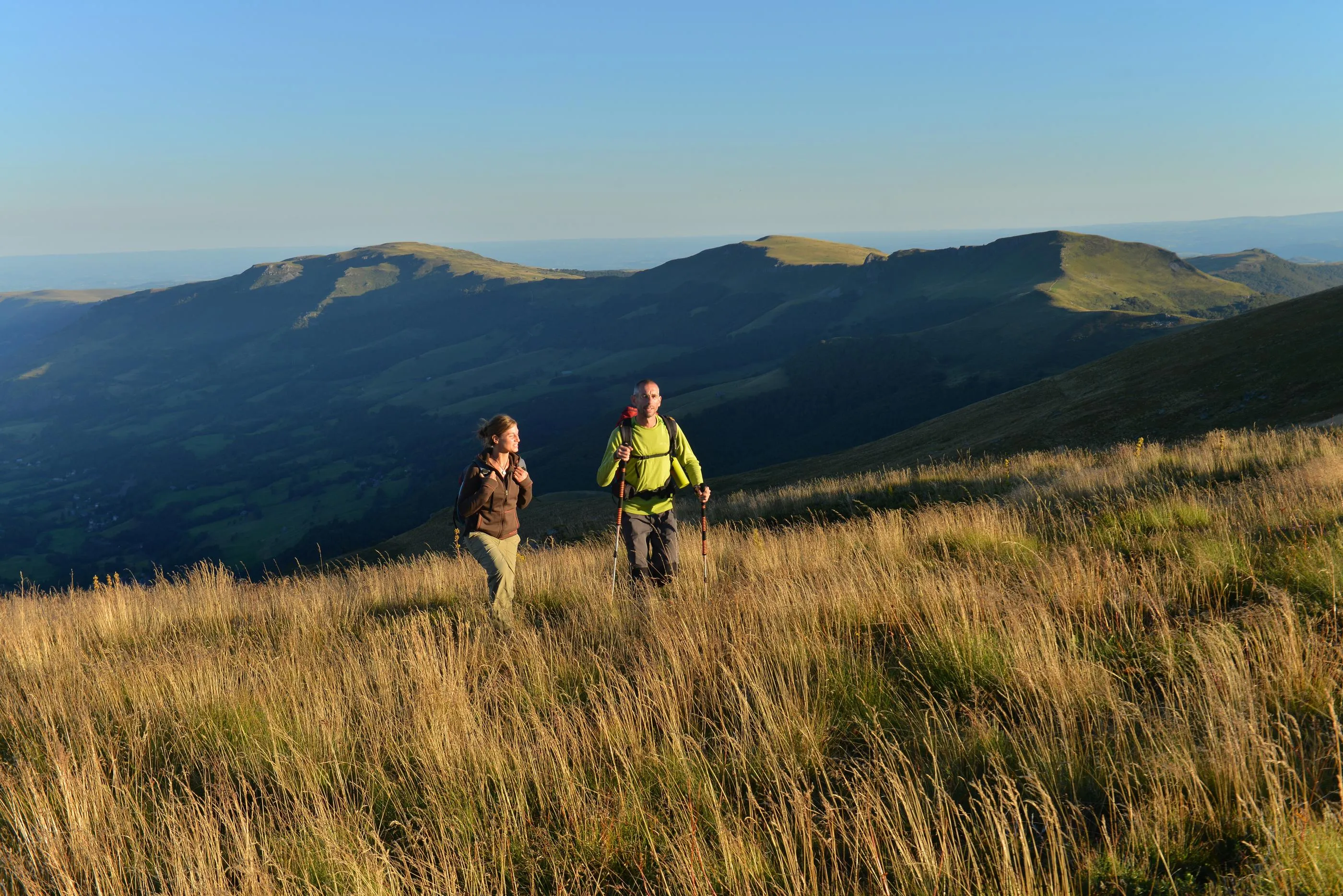 Deux personnes marchent dans un champ d'herbes hautes avec des montagnes en arrière-plan sous un ciel dégagé