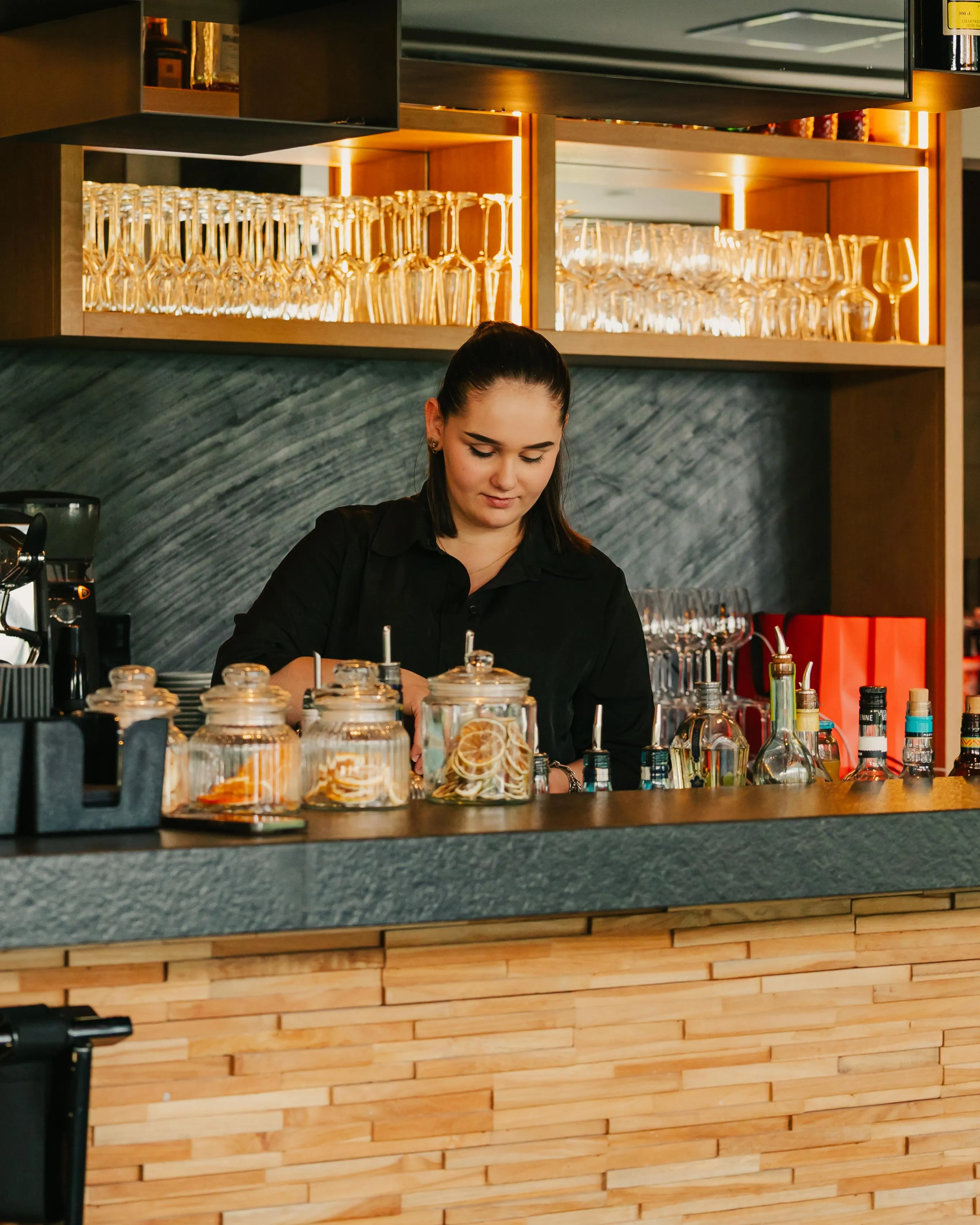 A woman prepares drinks behind a bar with glasses and bottles visible.