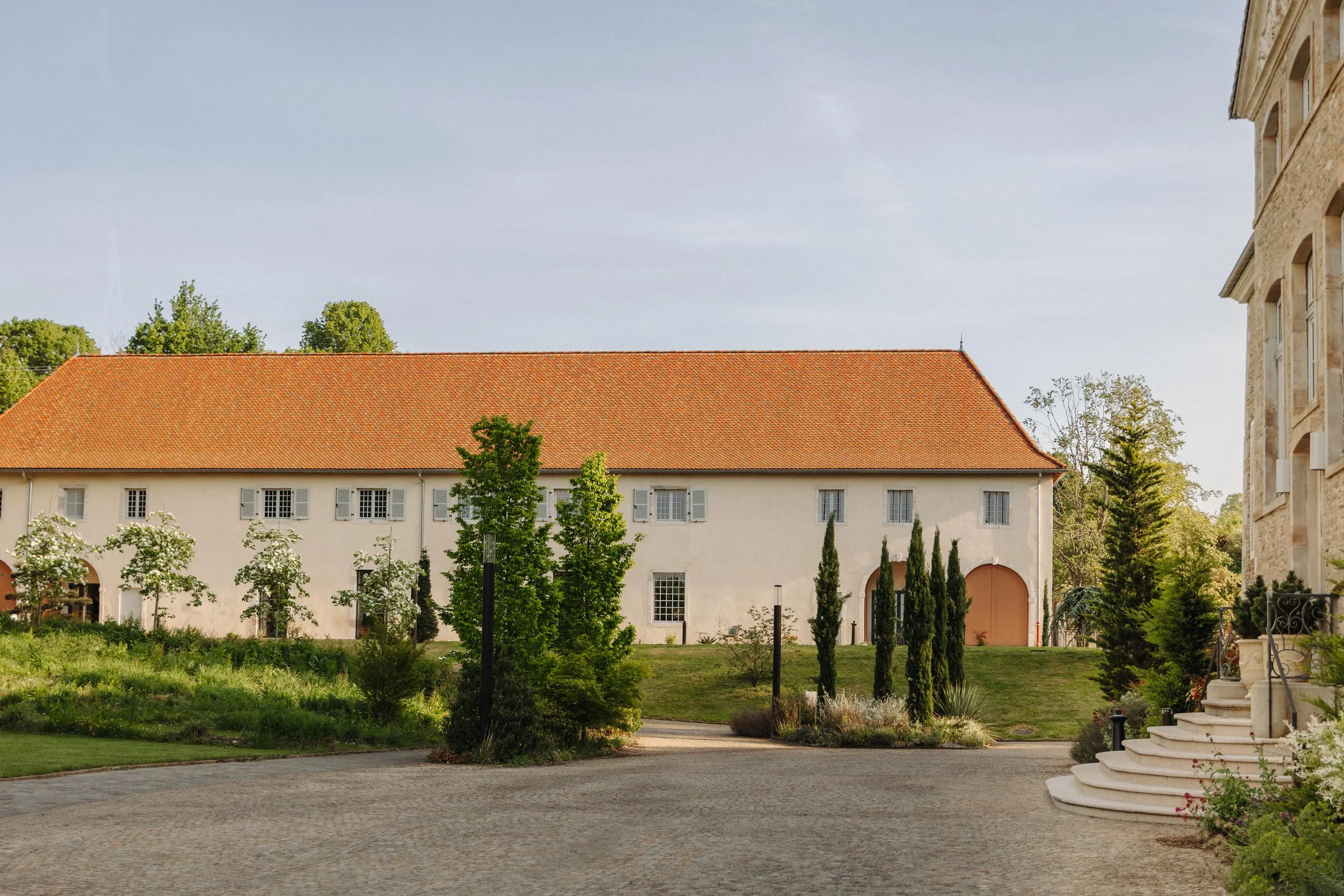 Ein Gebäude mit rotem Dach und grauen Fensterläden, umgeben von Bäumen und Vegetation, in einem gepflasterten Hof