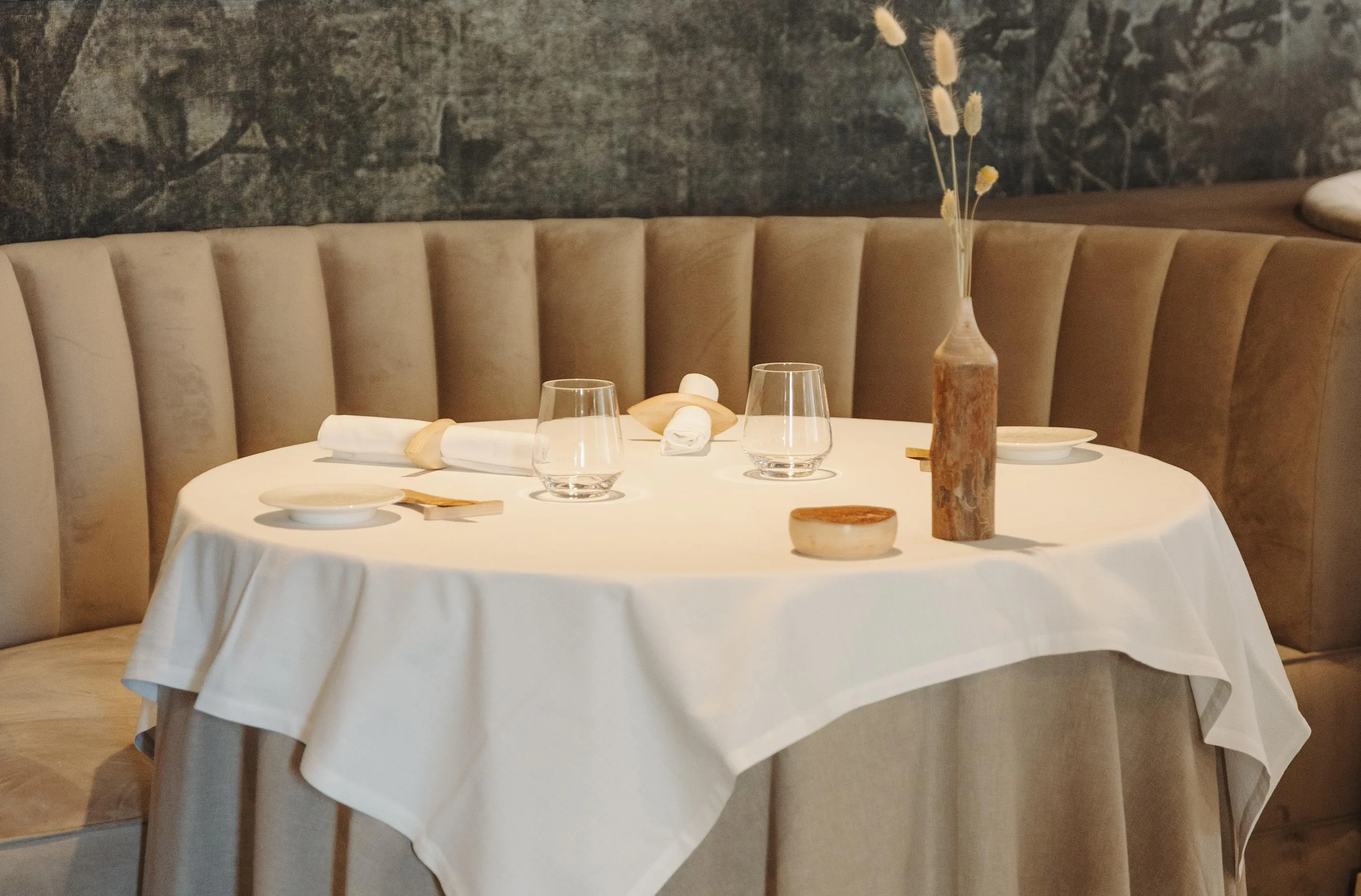 Table dressée avec verres, assiettes, serviettes et un vase contenant des fleurs séchées, entourée de banquettes en cuir beige