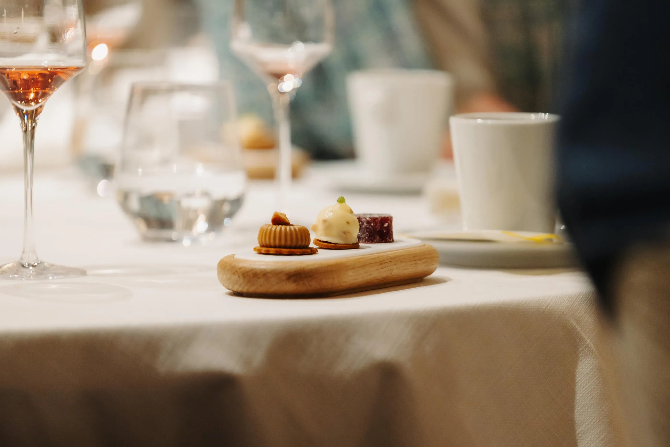Petite assiette en bois avec trois desserts et verres de vin à l'arrière-plan sur une table recouverte d'une nappe blanche
