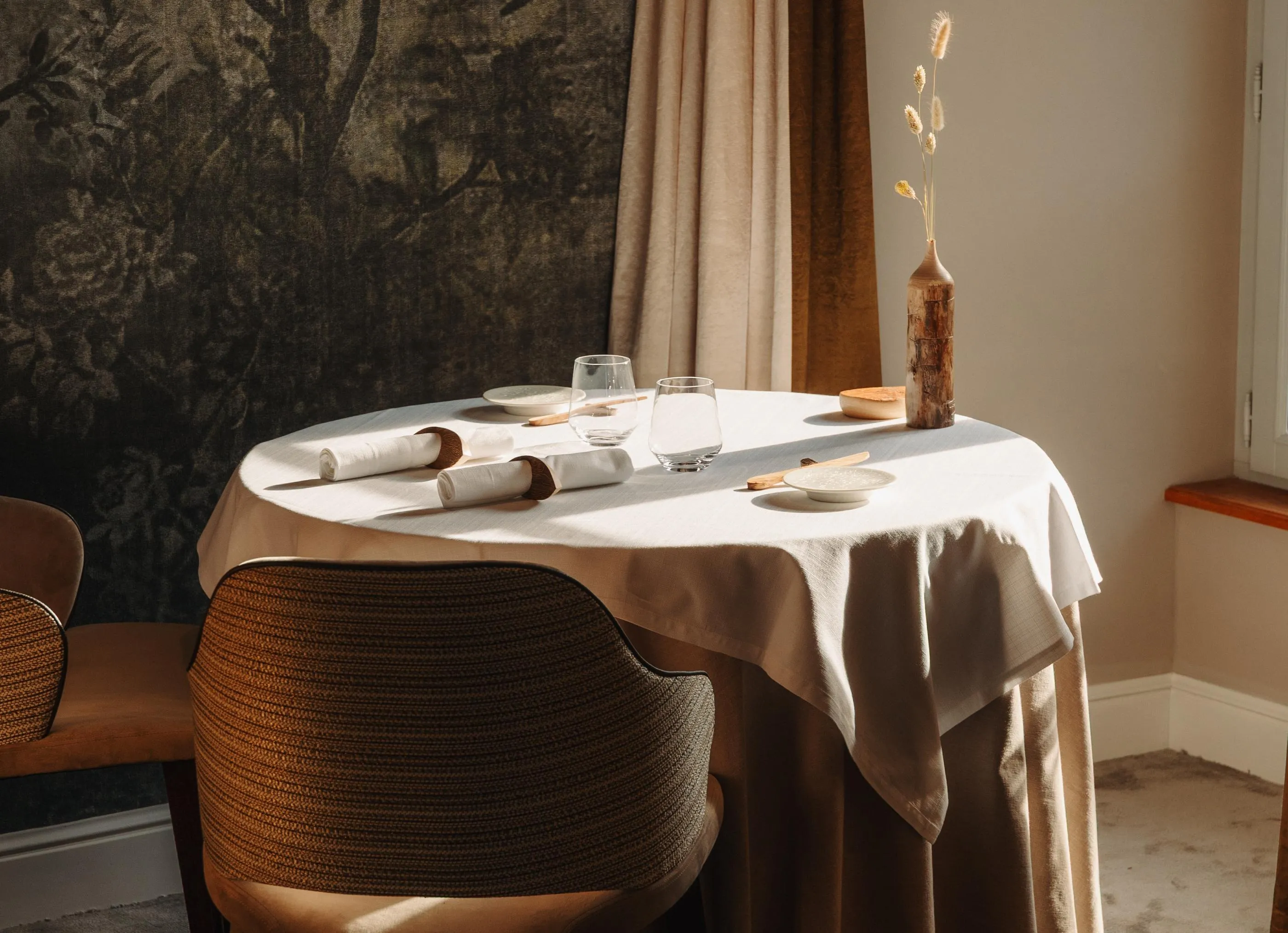 Table dressée avec des verres, des assiettes, des couverts et une décoration florale dans un cadre intérieur avec des chaises et des rideaux