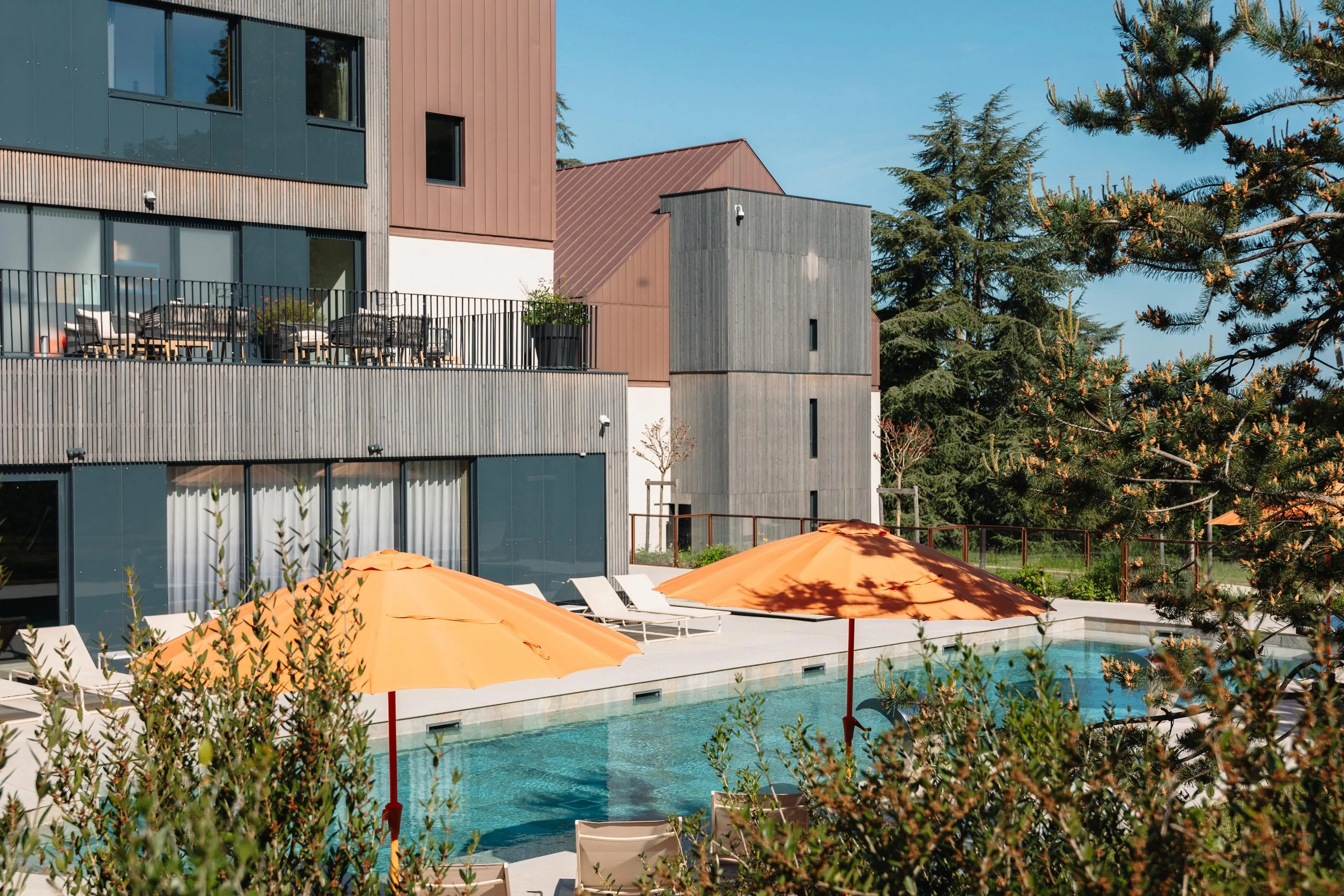 Außenpool mit Liegestühlen und orangen Sonnenschirmen vor einem modernen Gebäude mit Holz- und Metallfassaden