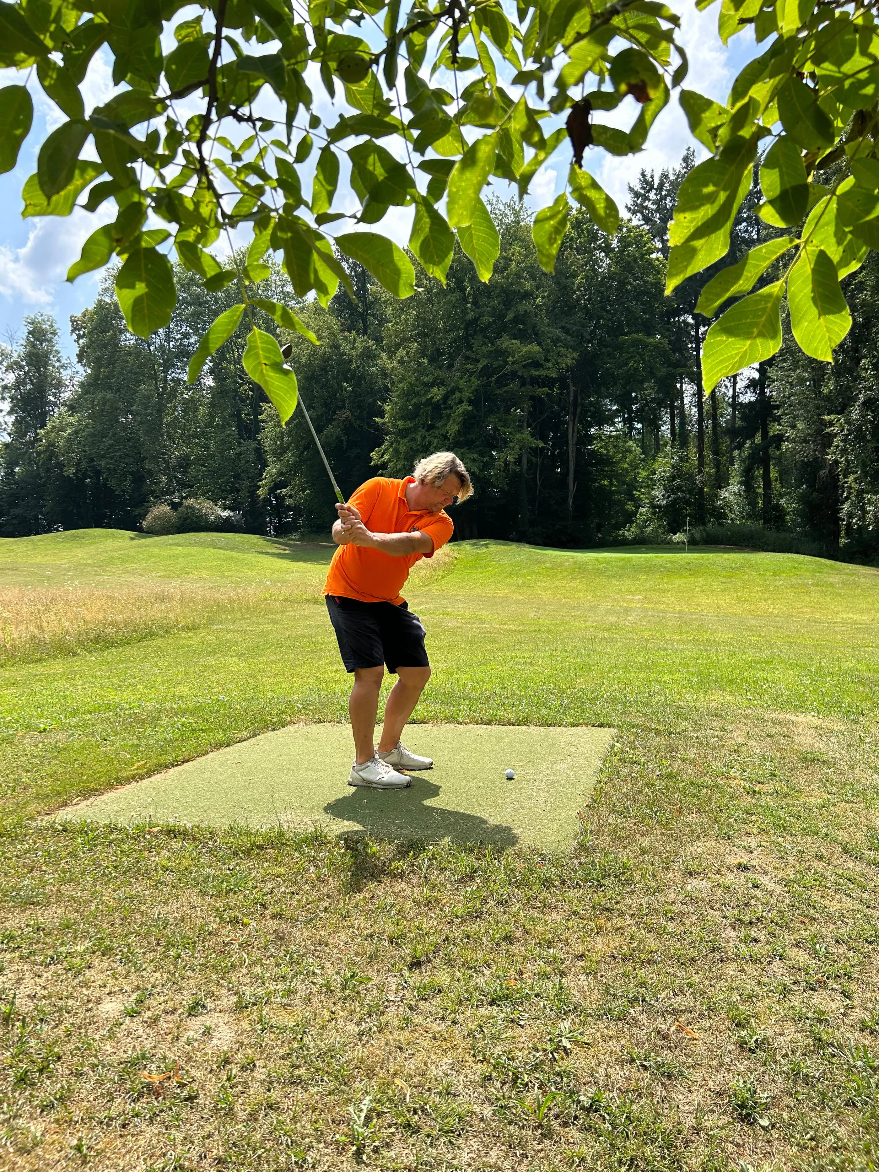 Une personne en tenue orange et noire joue au golf sur un green bordé d'arbres et d'herbe sèche