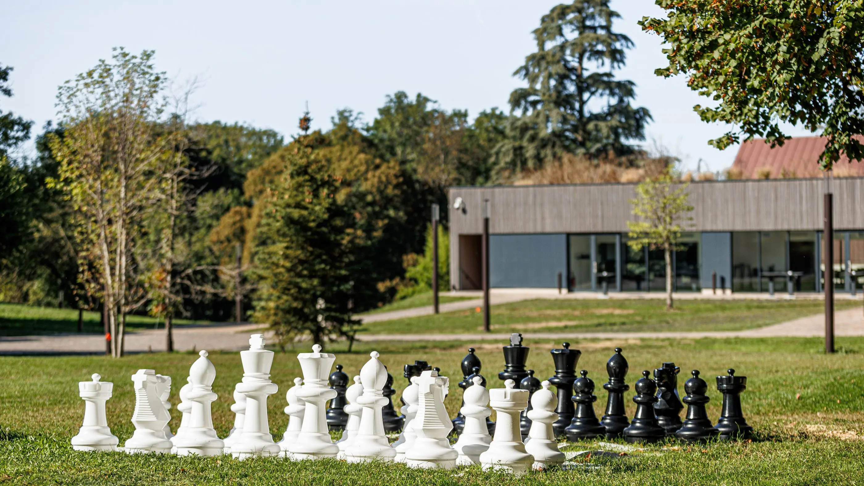 Un jeu d'échecs géant en plein air avec des pièces blanches et noires disposées sur l'herbe devant un bâtiment moderne entouré d'arbres