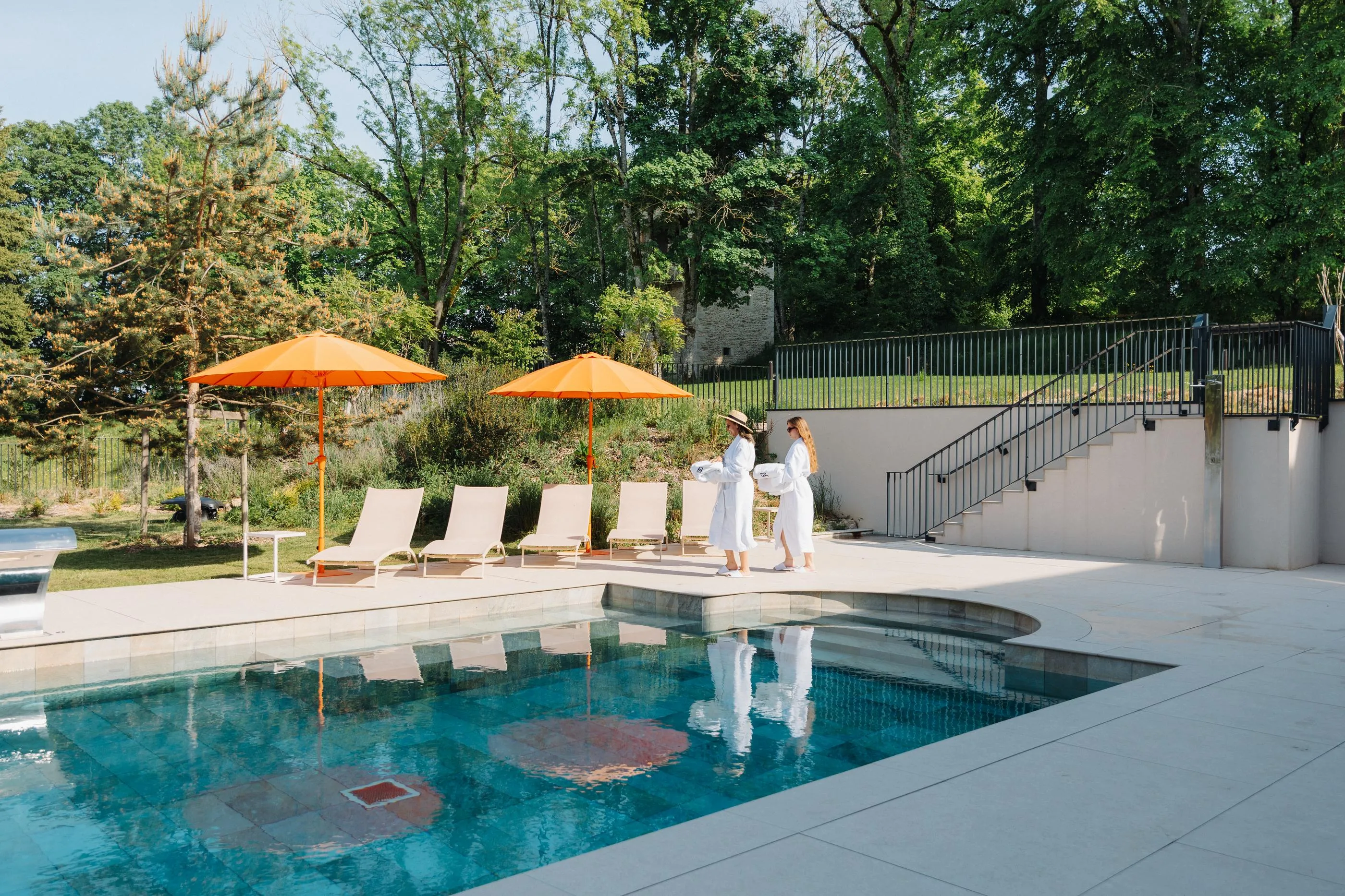 Two people in white walk near a pool surrounded by sun loungers and orange umbrellas, in a tree-lined garden.