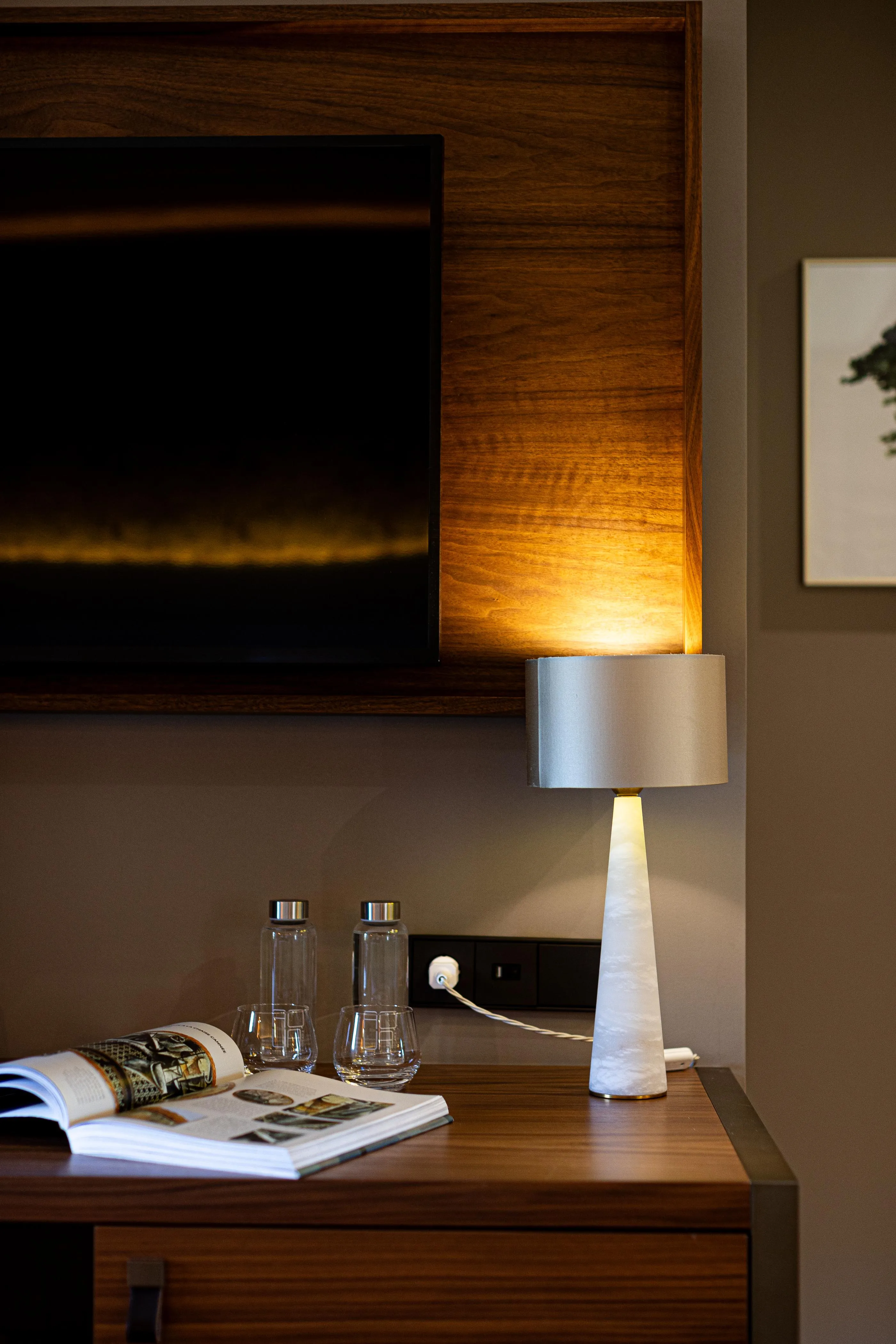 A wooden nightstand with a lit lamp, glasses, and magazines.