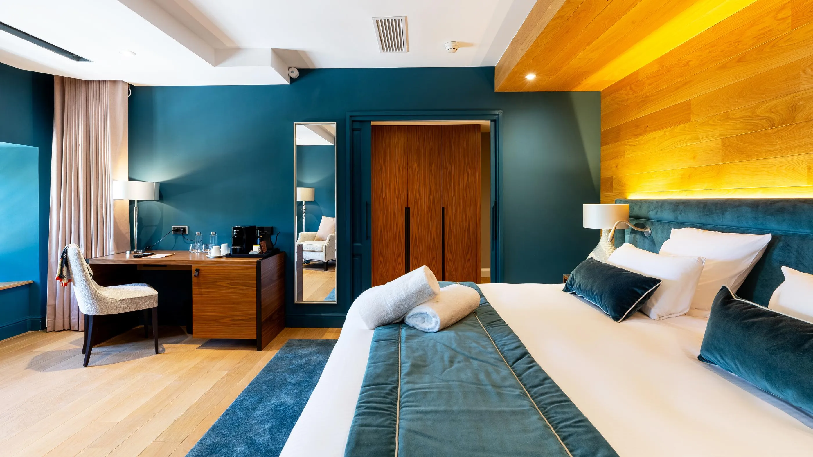 Hotel room with double bed, desk, chair, and wooden door, blue-painted walls, and light wooden ceiling.