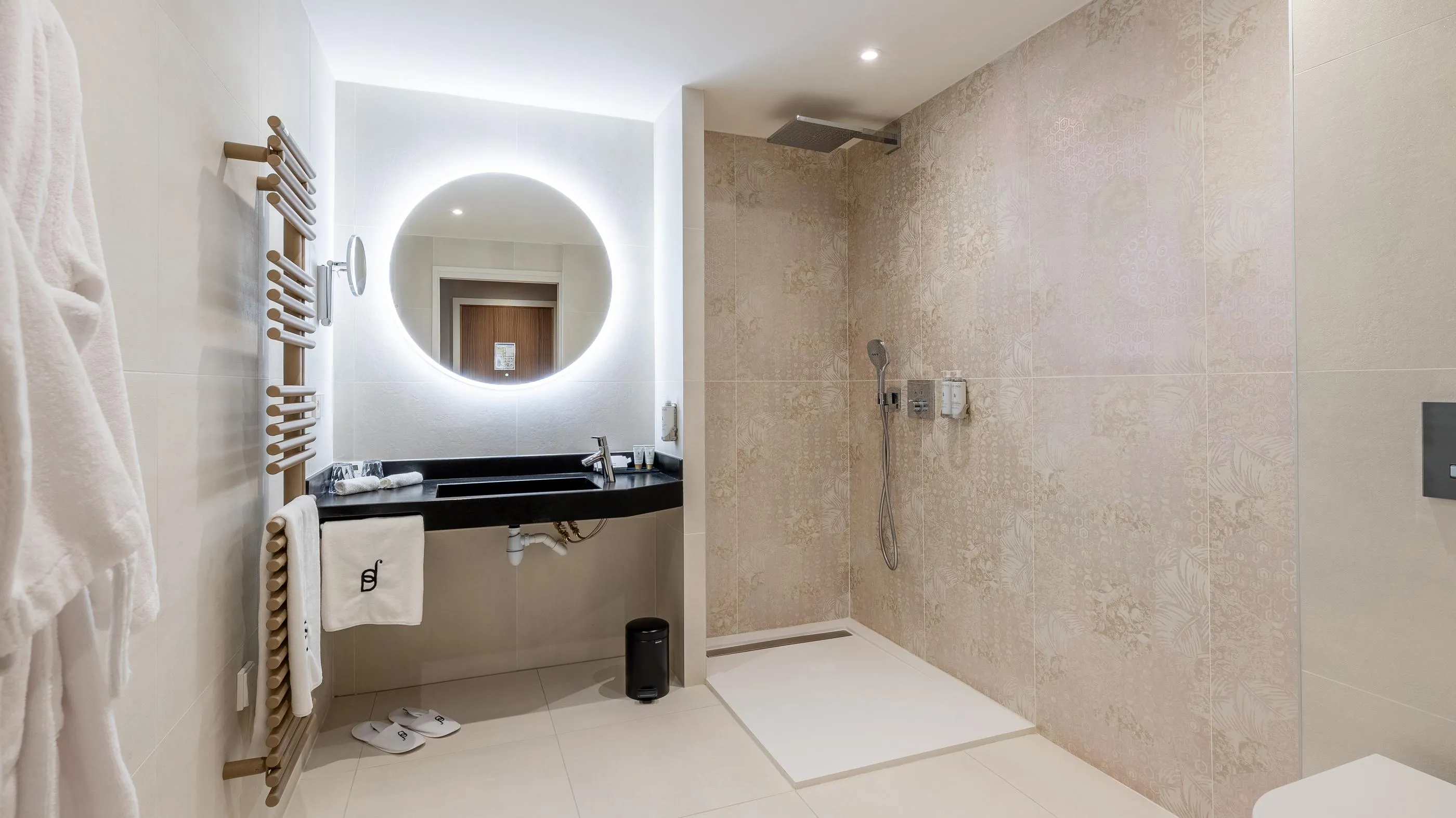 Modern bathroom with sink, illuminated round mirror, walk-in shower, and hanging towels