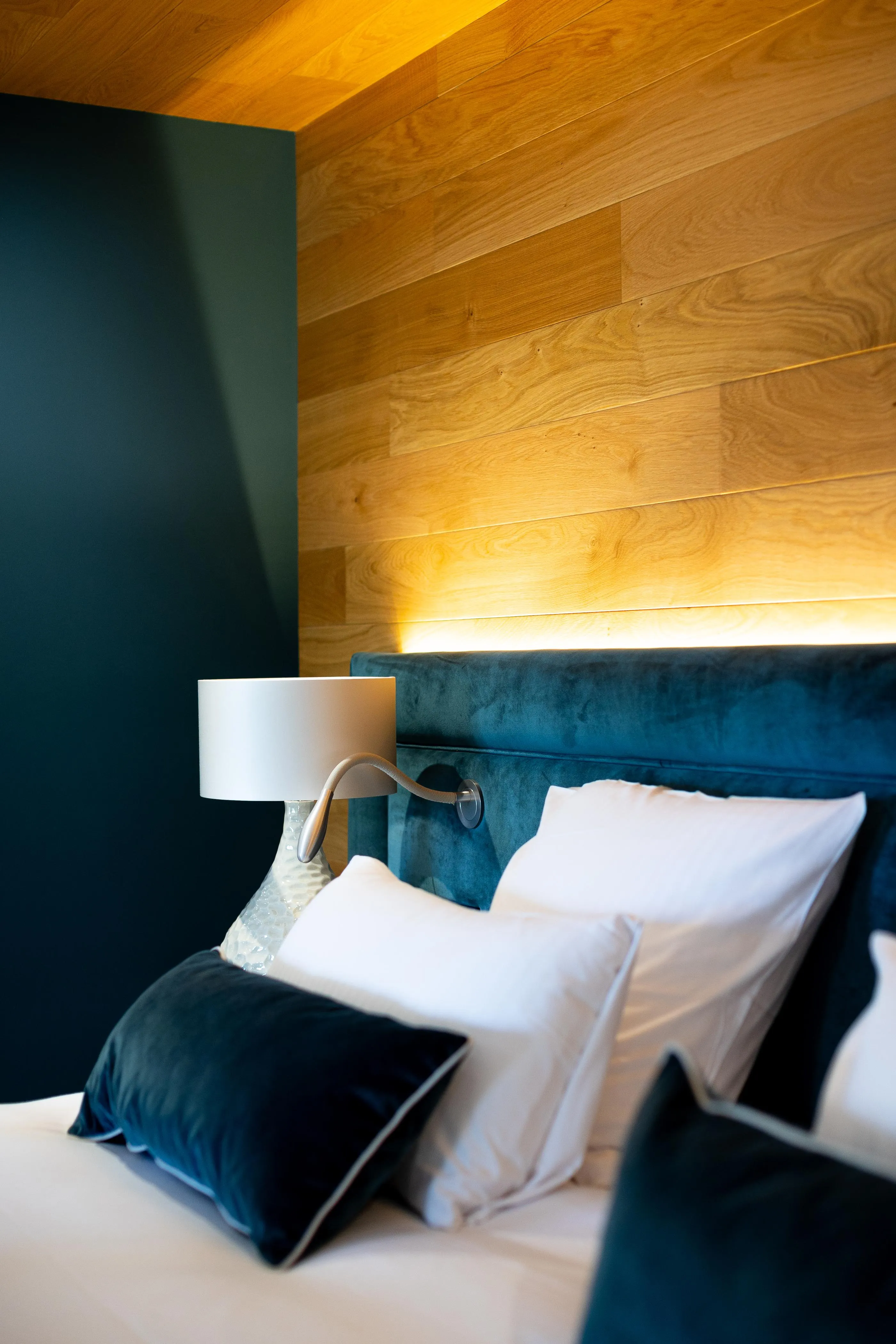 Bed with a dark green fabric headboard, white and black pillows, a bedside lamp, and a wood-paneled wall lit in the background.