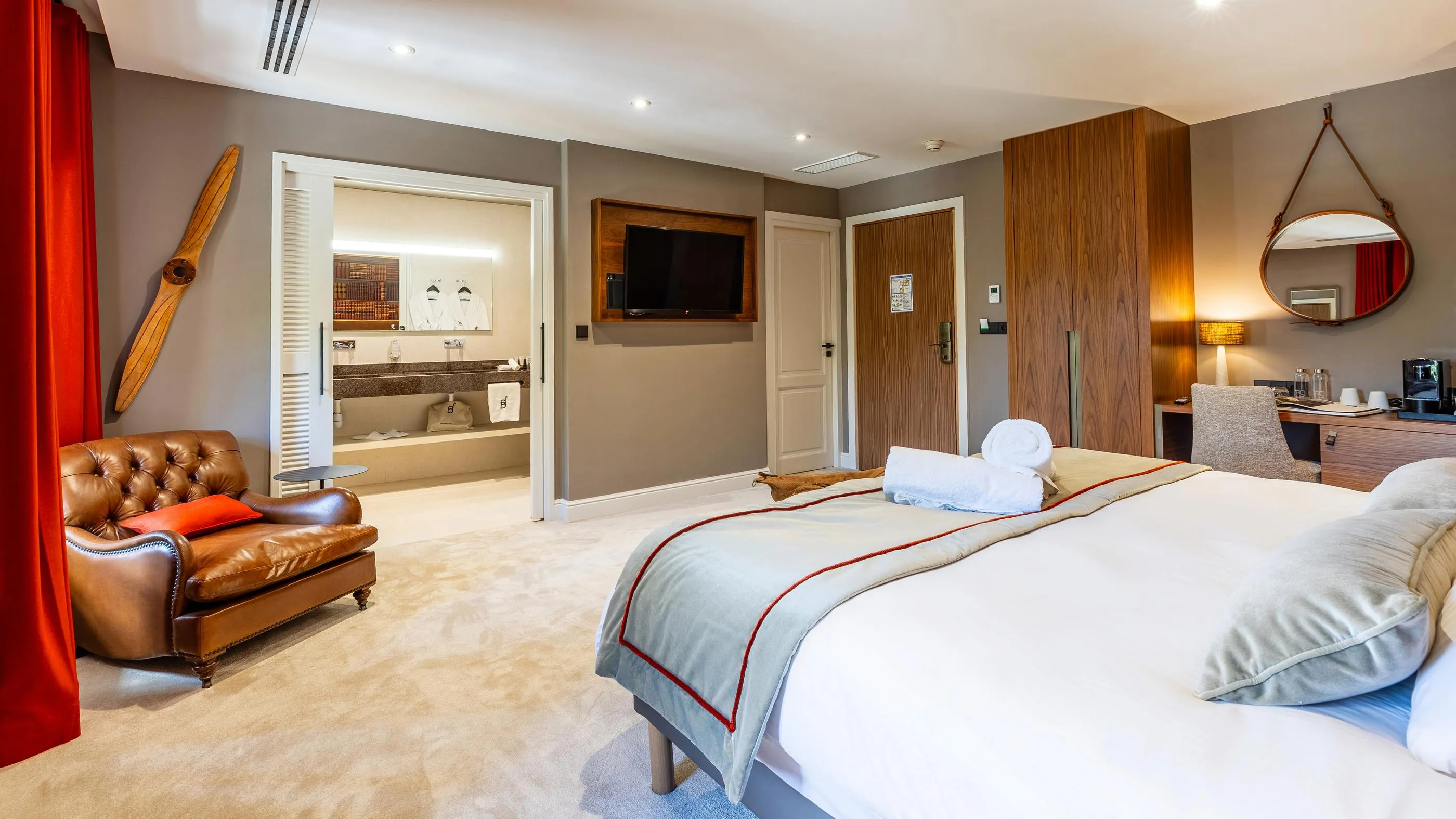 Hotel room with double bed, leather armchair, television, mirror, and bathroom visible in the background