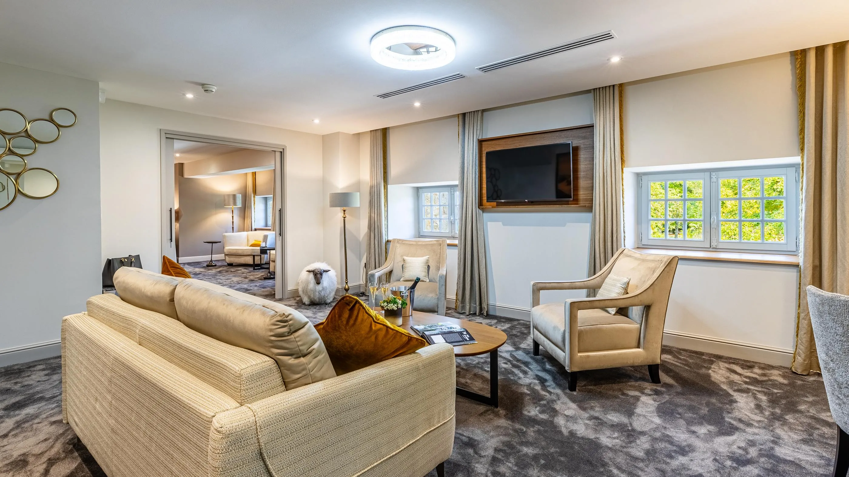 Living room with beige sofa, armchairs, coffee table, and television, view of another room with bed