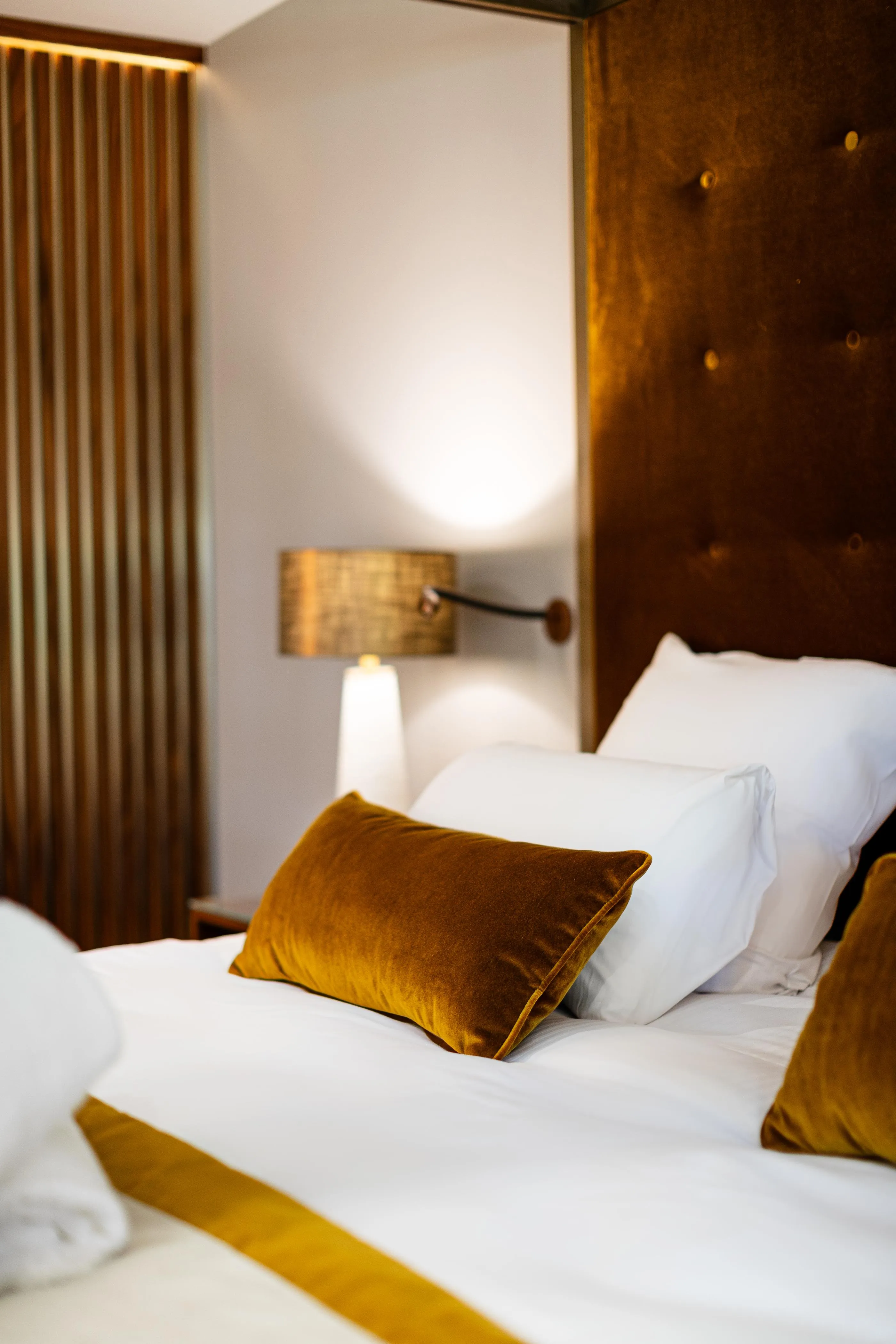 Bed with white sheets, gold velvet cushions, and a lit bedside lamp