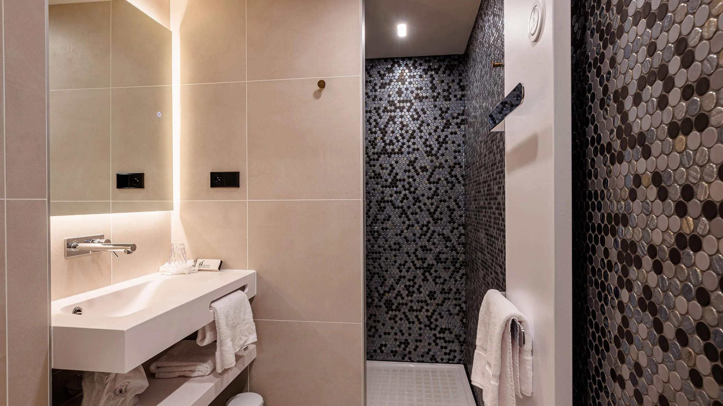 Salle de bain avec lavabo, miroir éclairé, serviettes et douche aux murs recouverts de mosaïque sombre