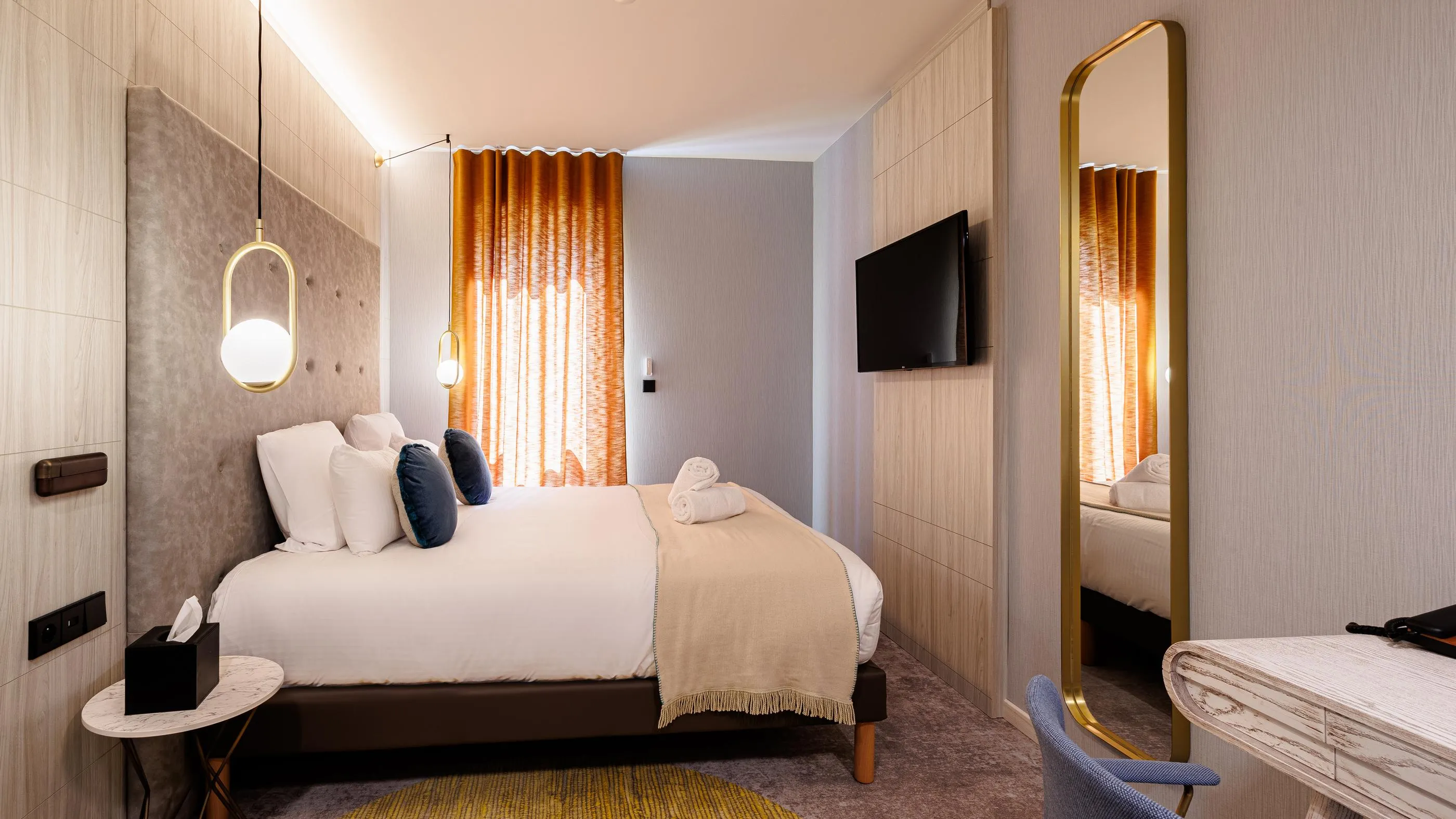 Hotelzimmer mit einem Doppelbett, einem Wandfernseher, hängenden Leuchten und einem Fenster mit goldenen Vorhängen