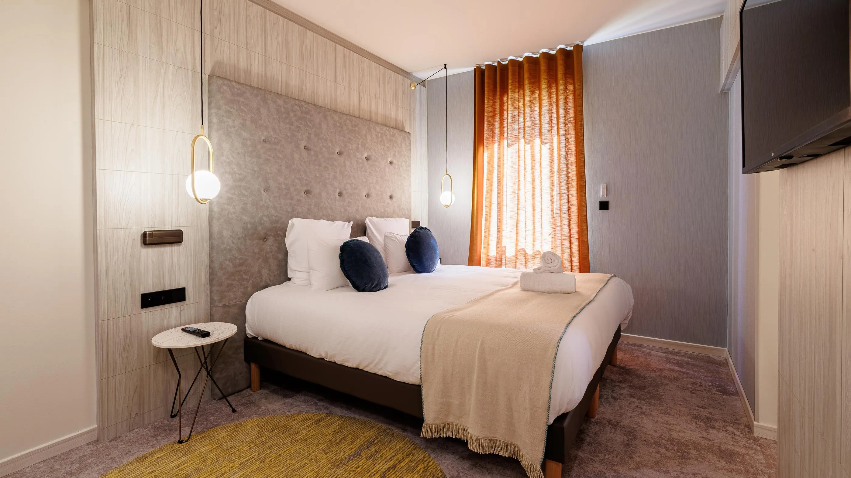 Chambre avec un lit double recouvert d'une couverture beige, deux coussins bleus et une table de chevet avec une lampe suspendue