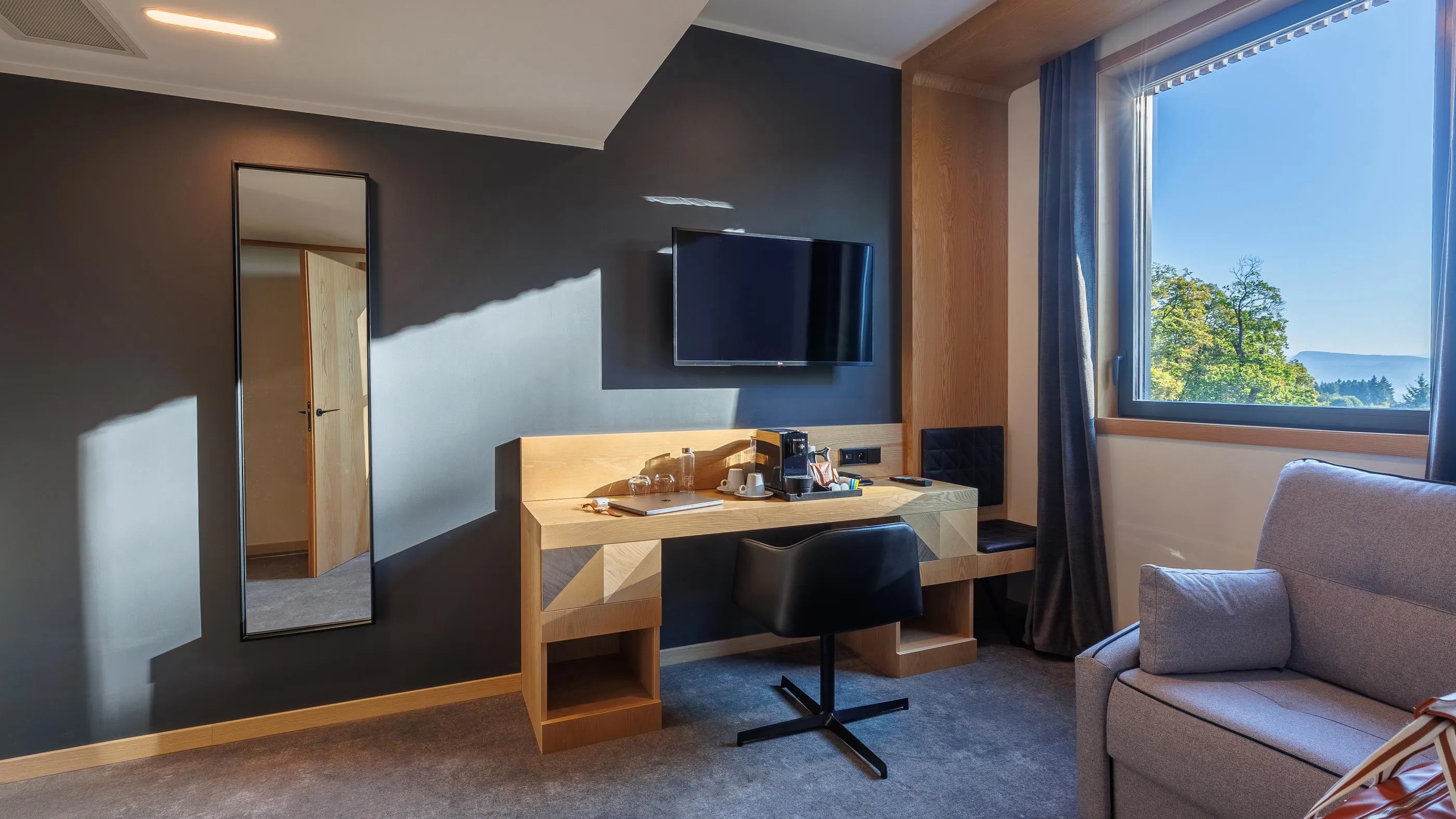 Zimmer mit Holzschreibtisch, schwarzem Stuhl, Wandfernseher, Fenster mit Naturblick und grauem Sofa