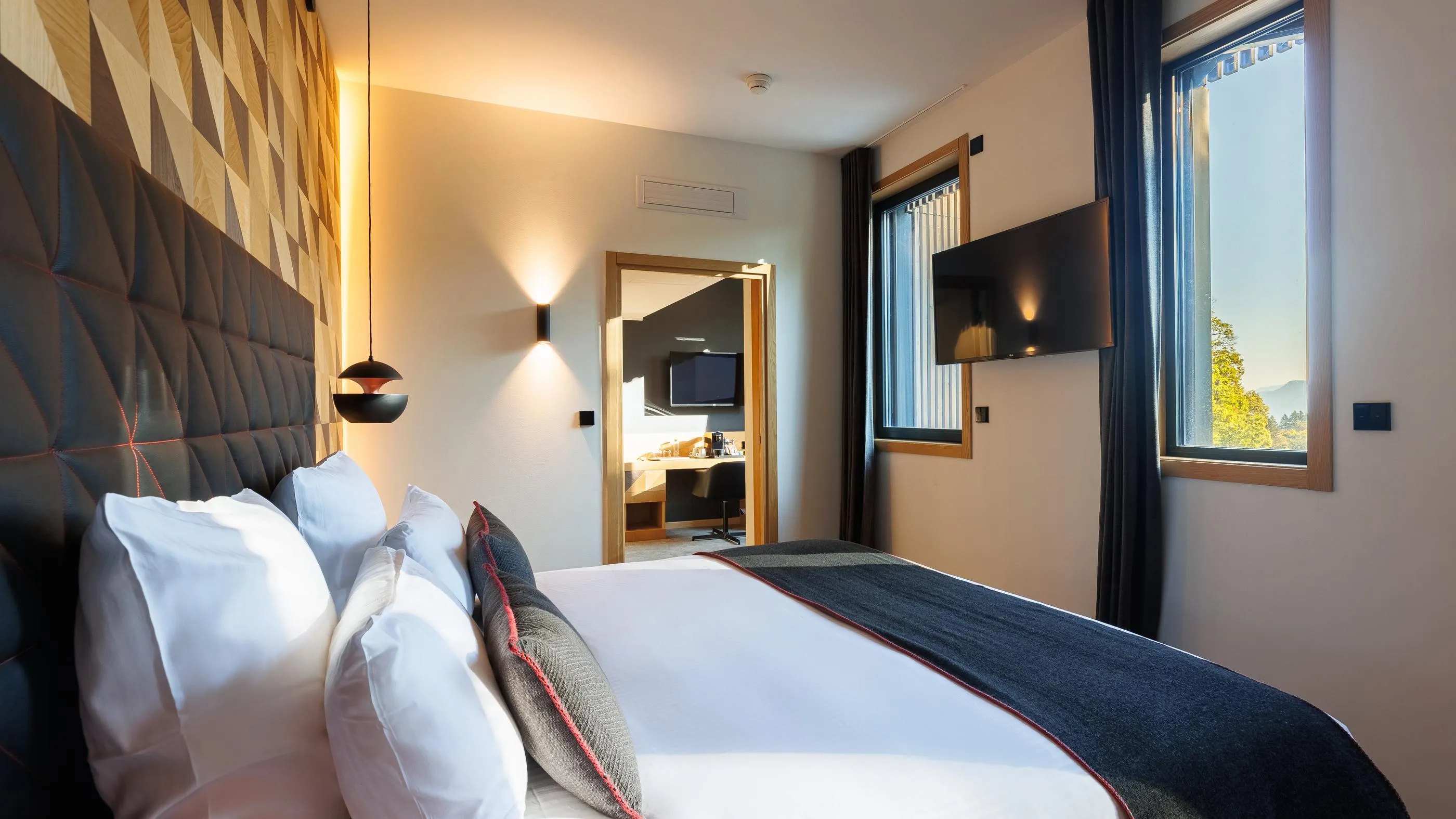 Hotelzimmer mit einem Doppelbett, einer gepolsterten Kopflehne und Kissen, einem Wandfernseher und einem Fenster mit Aussicht nach außen