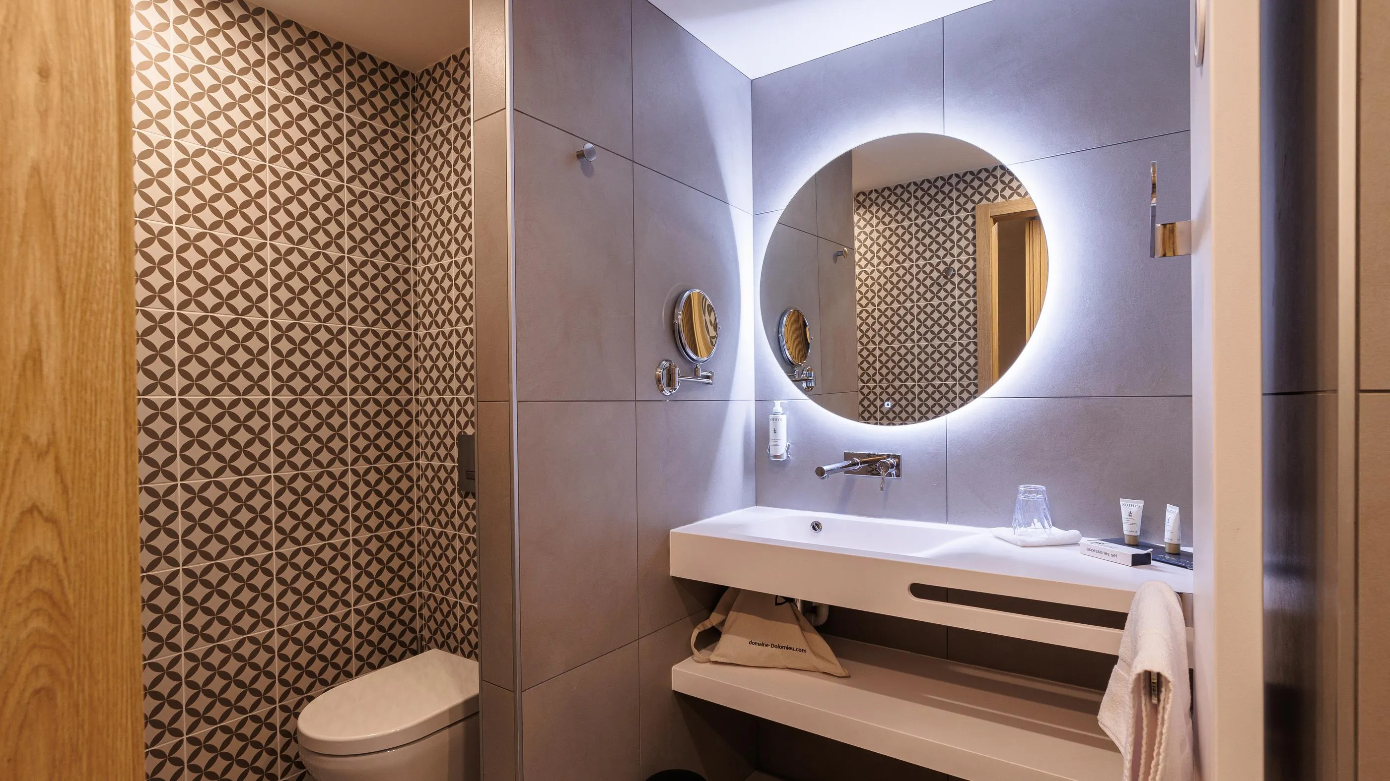 Modern bathroom with sink, illuminated round mirror, toilet, and geometrically patterned tiled wall